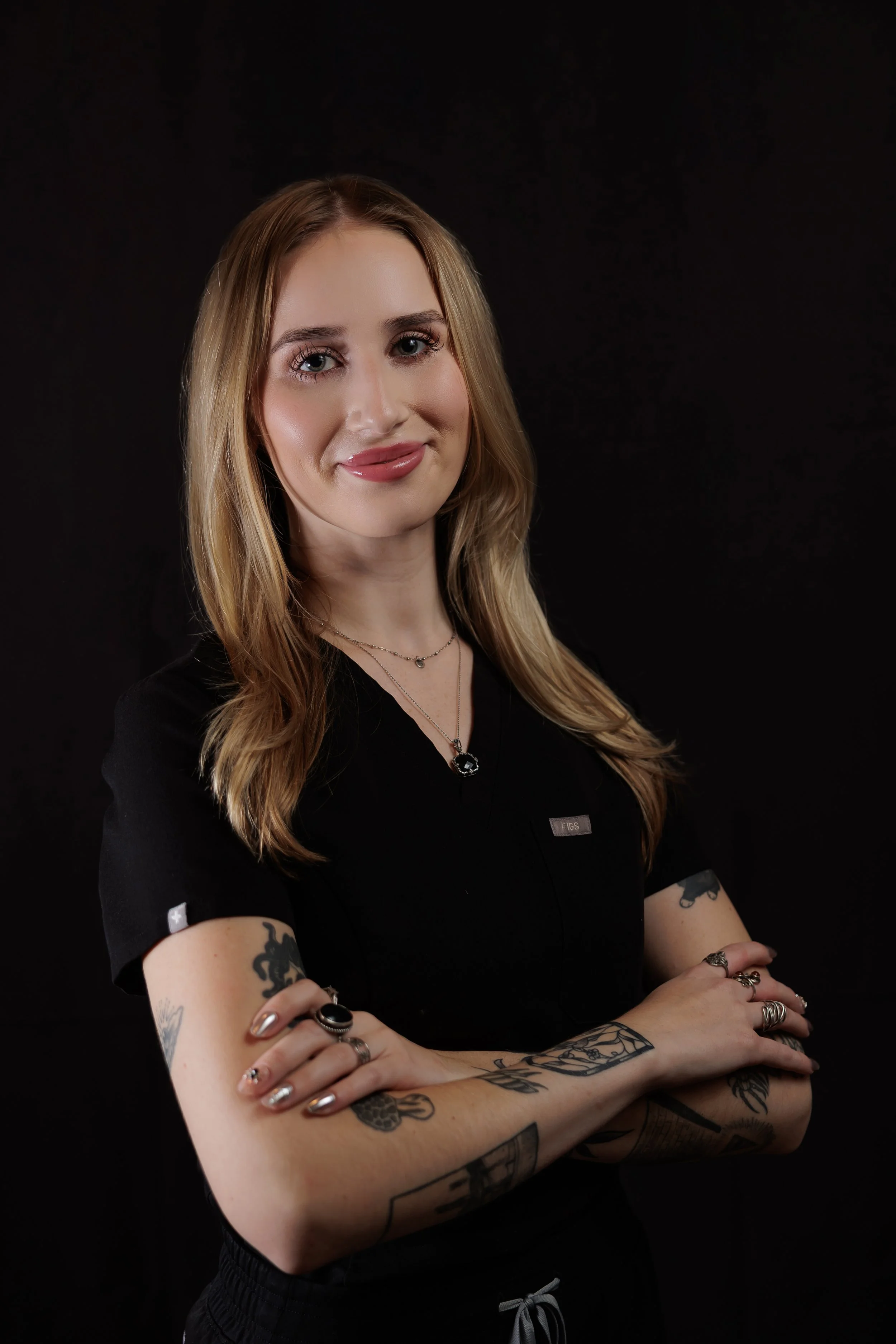 A young woman with long, wavy blonde hair and tattoos on her arms, wearing a black shirt, standing against a black background.