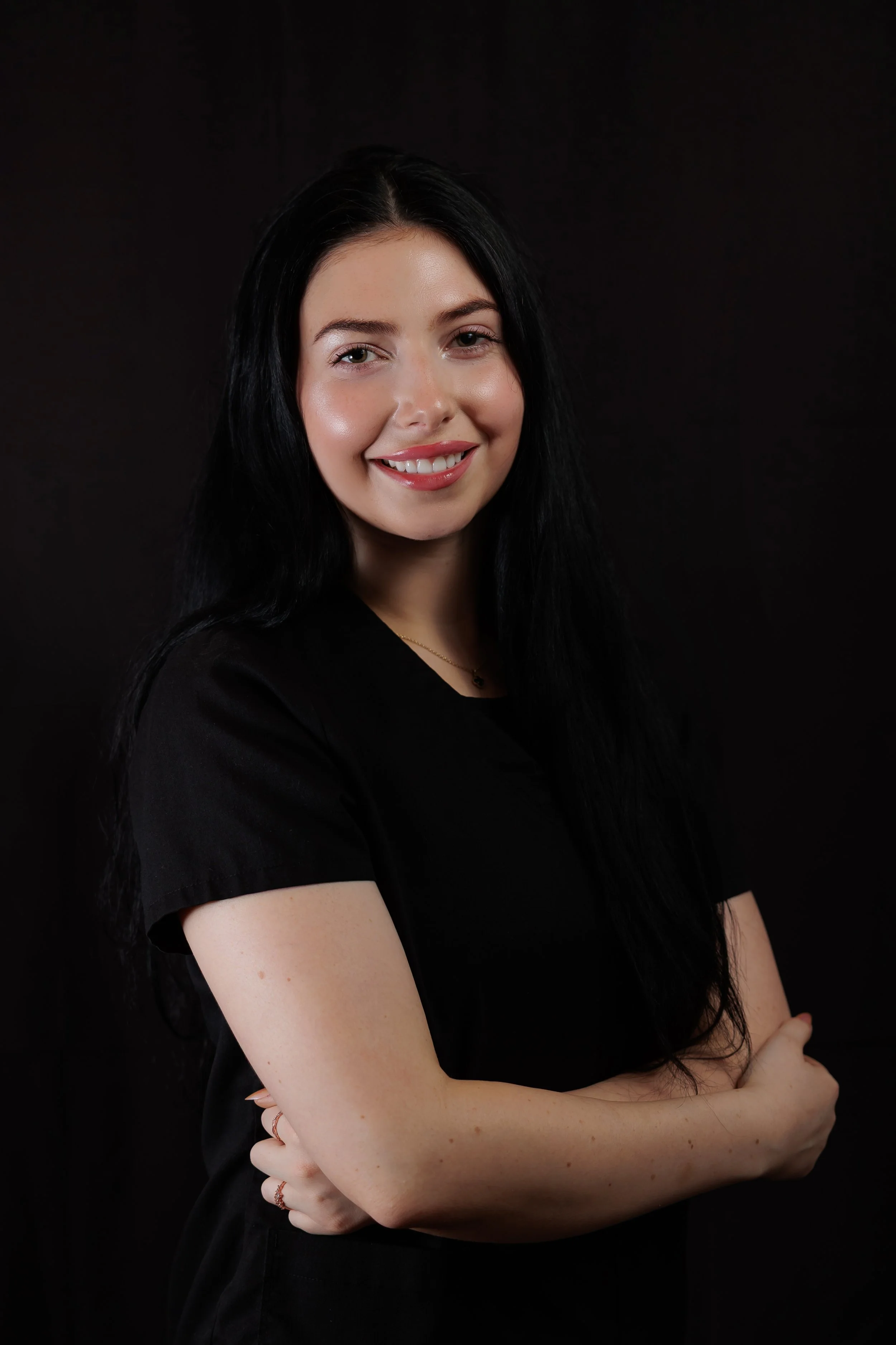 A portrait of a woman with long black hair, smiling, wearing a black top, with arms crossed in front of a dark background.