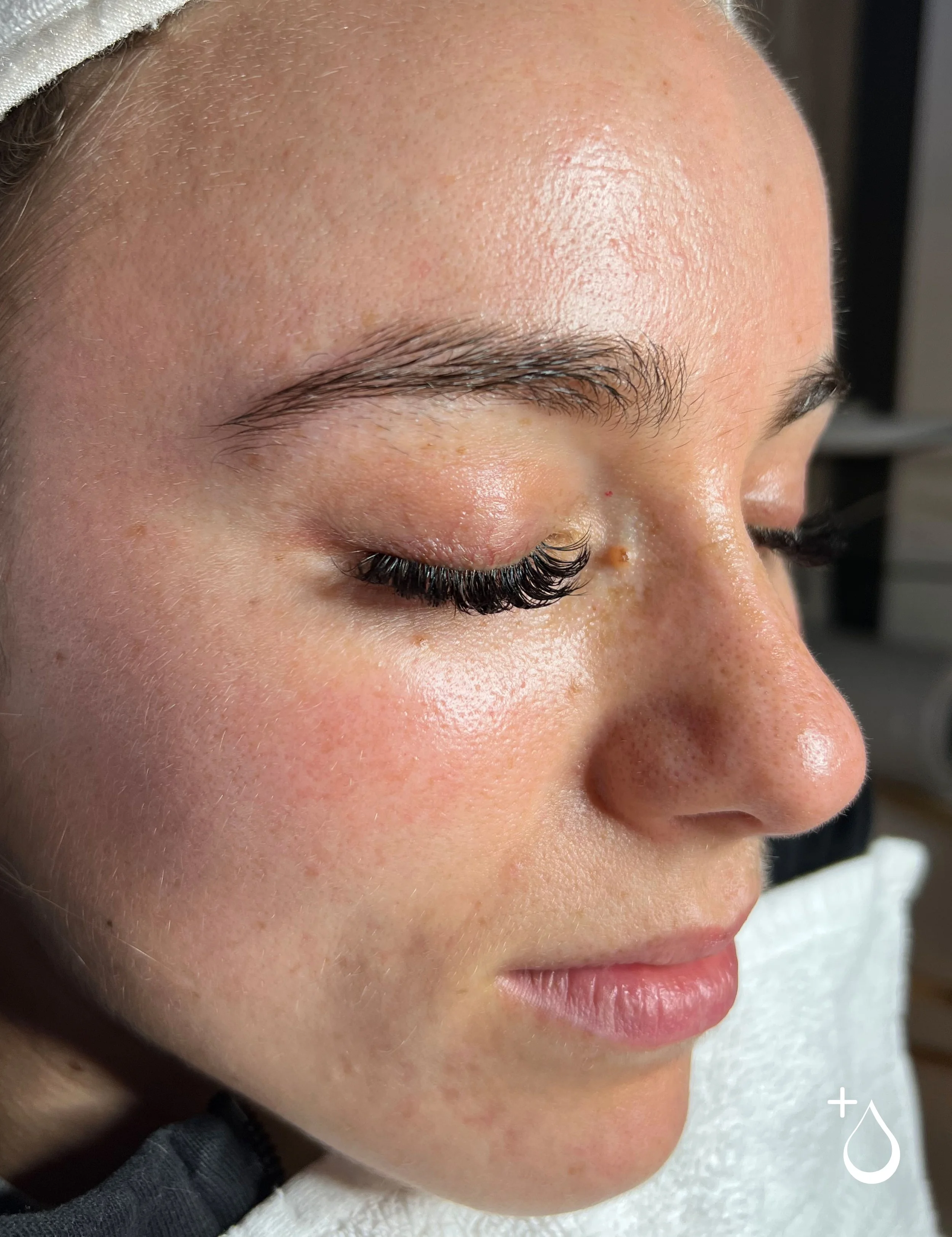 Close-up side view of a woman with eyelash extensions, her eyes closed, showing smooth skin with some freckles, and natural eyebrows.