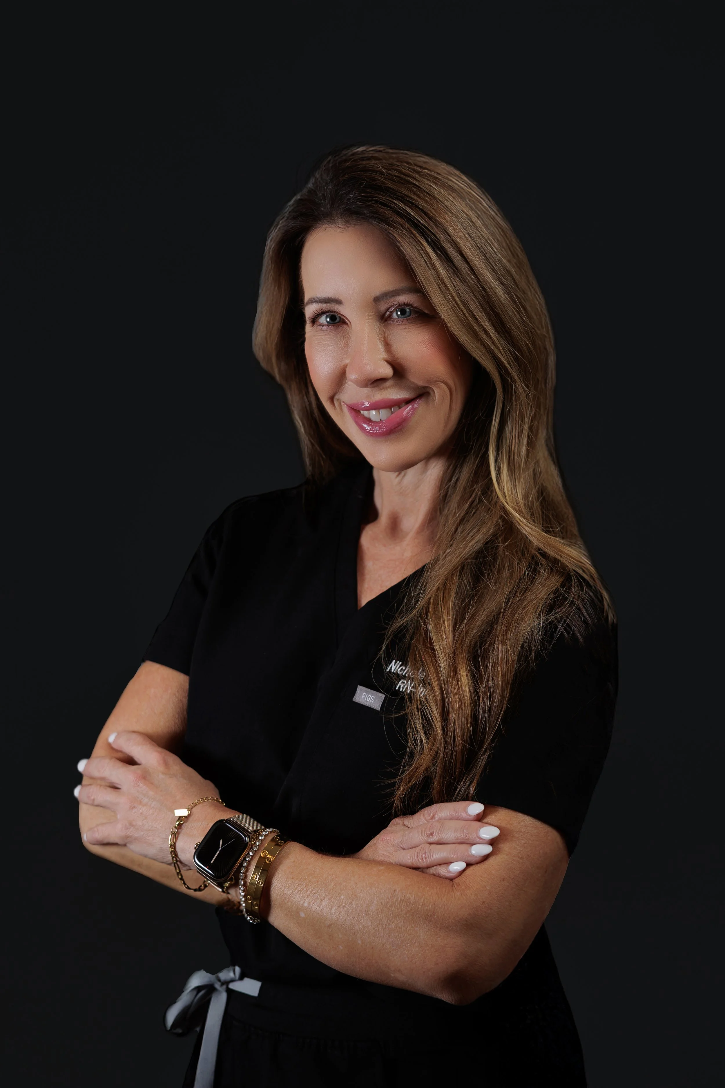 A woman with light skin and long, wavy brown hair, smiling and posing with arms crossed, wearing a black medical uniform with a name badge against a dark background.
