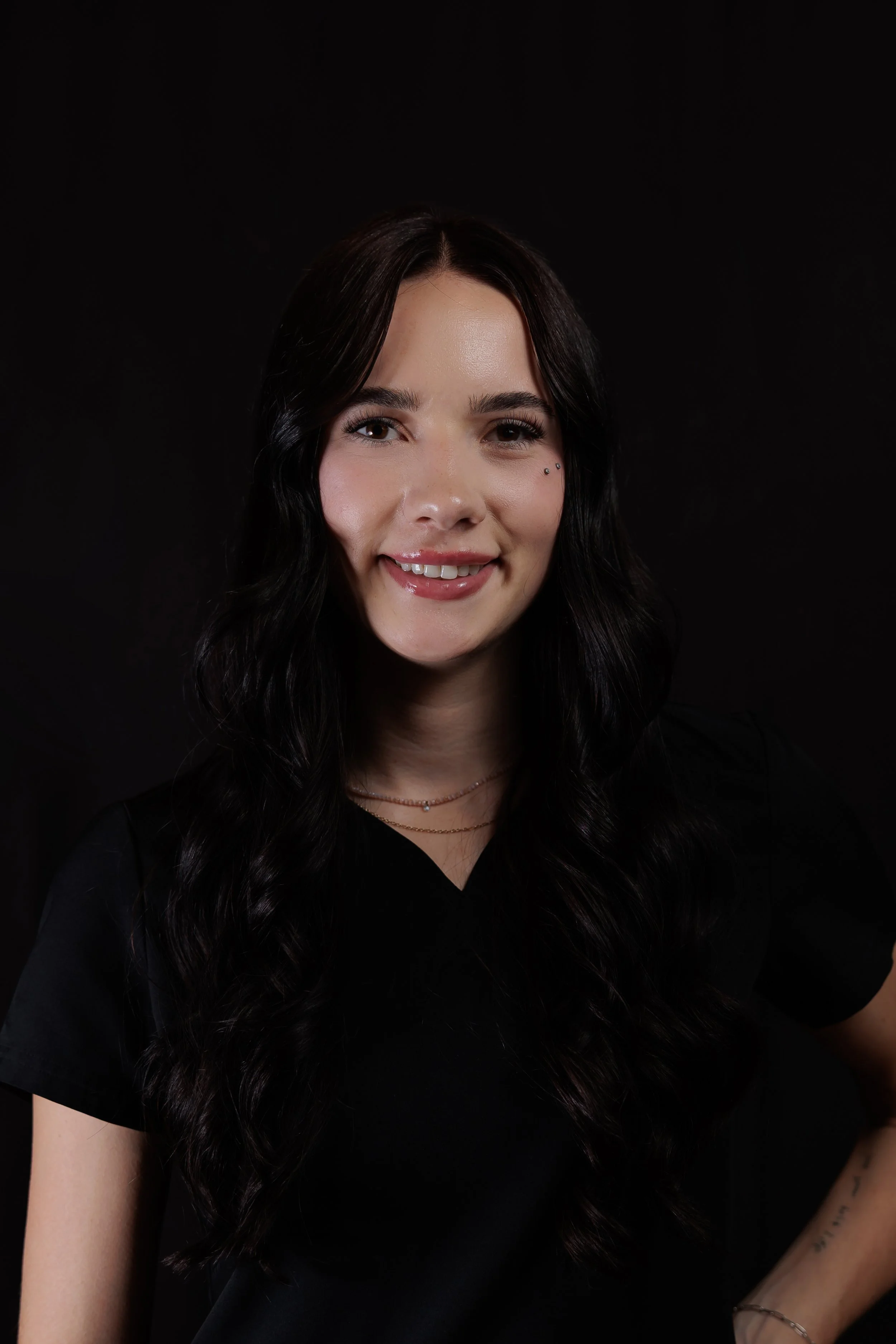 Portrait of a young woman with long, wavy black hair, smiling, wearing a black top, jewelry, and a small facial piercing, against a black background.