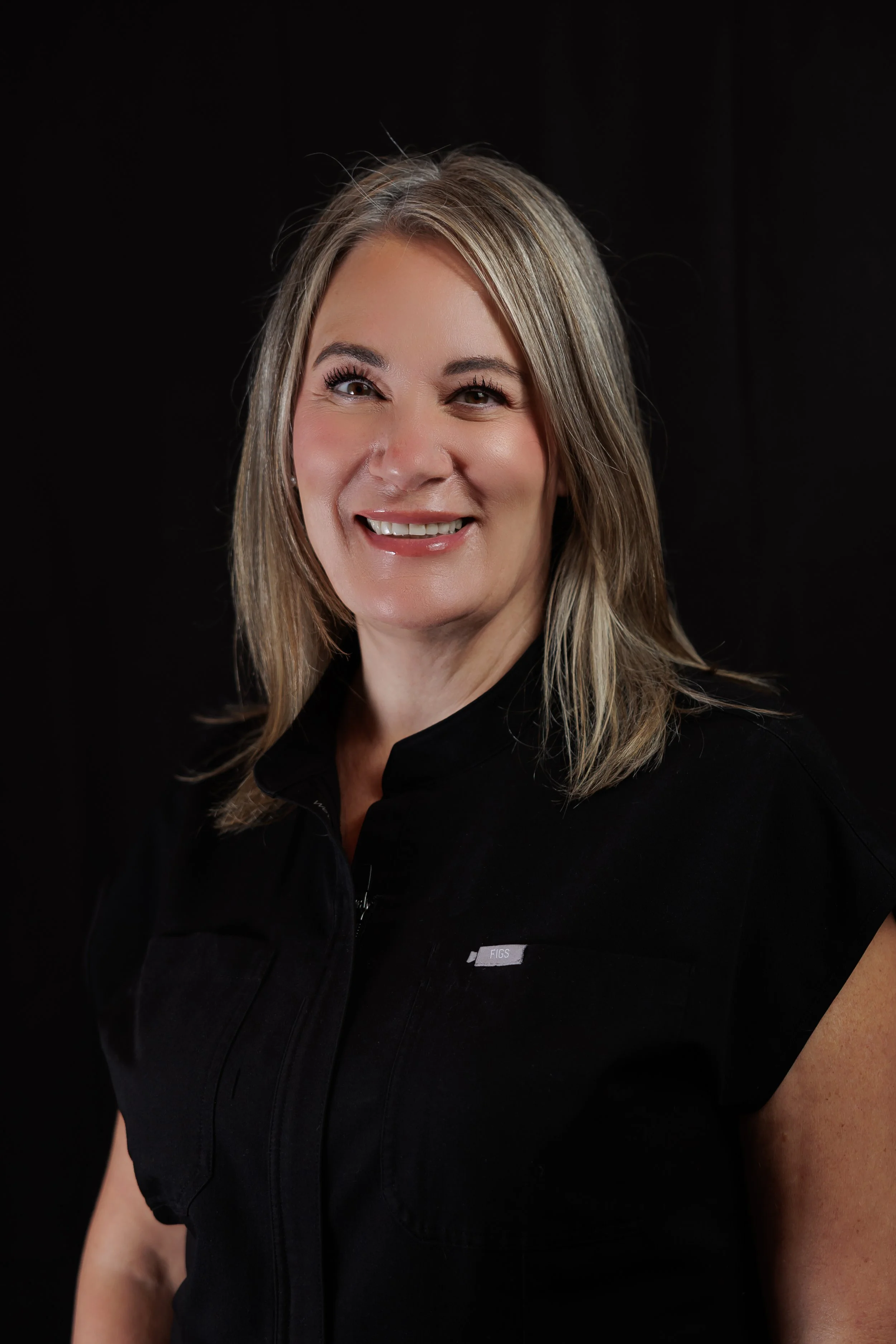 A smiling woman with shoulder-length blonde hair wearing a black vest against a dark background.