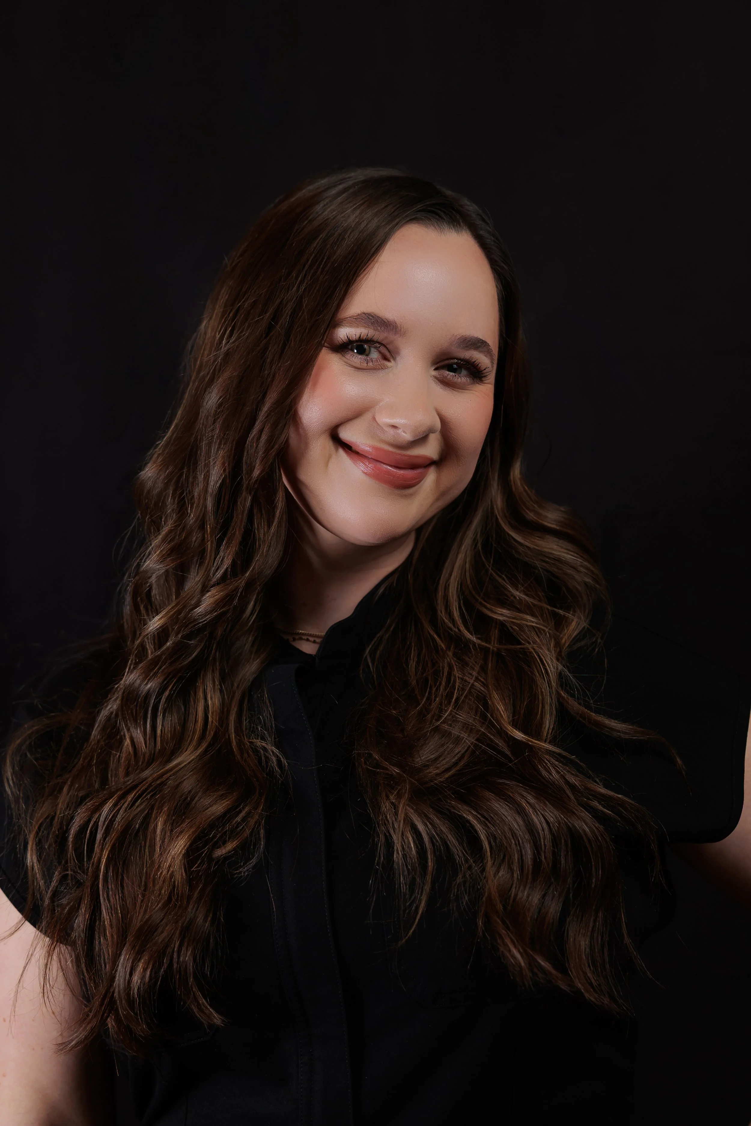 Portrait of a smiling woman with long wavy brown hair, wearing a black top, against a dark background.