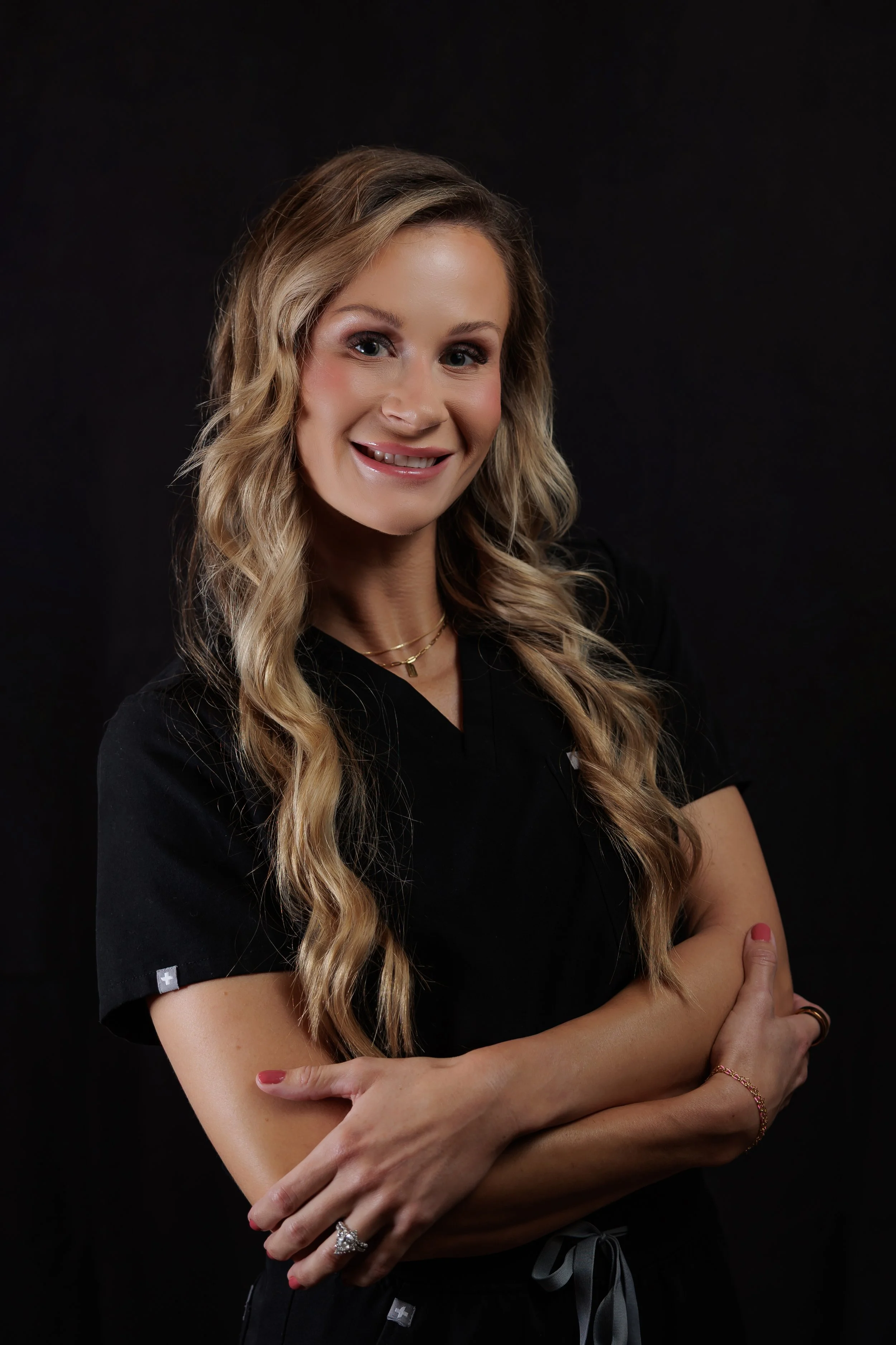 A woman with long blonde curly hair smiling, wearing black scrubs and jewelry, crossing her arms, against a black background.