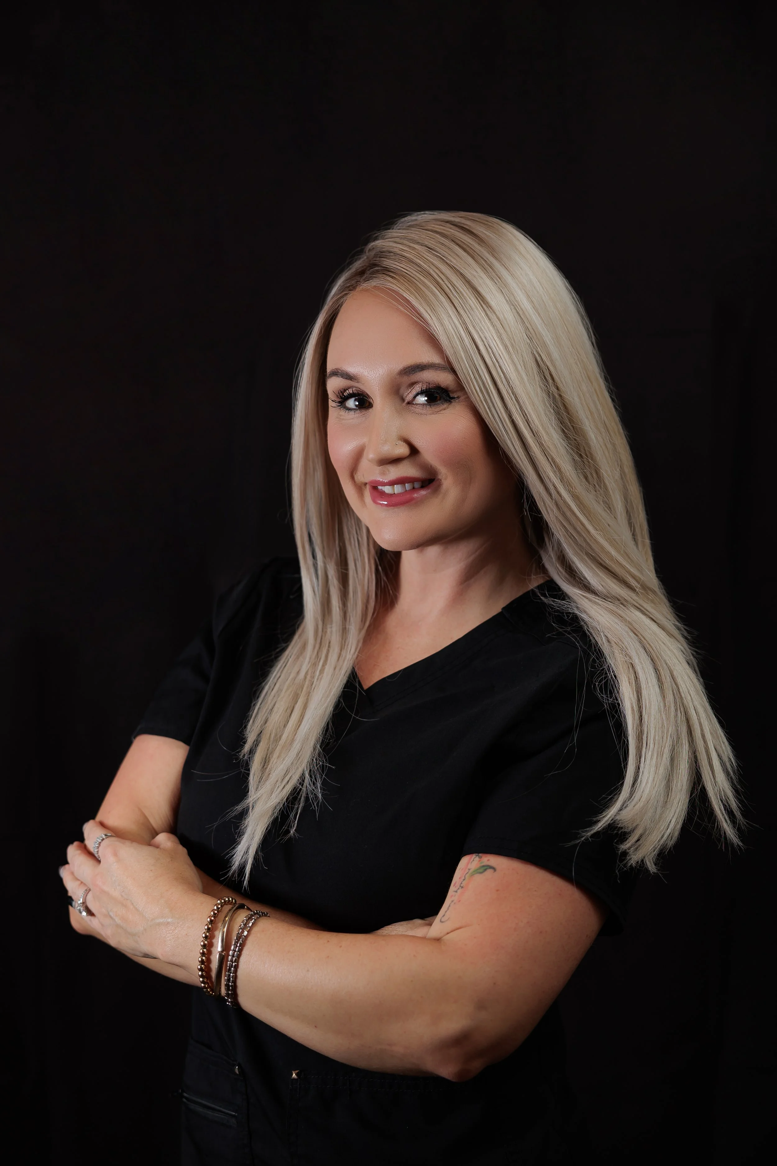 Portrait of a smiling blonde woman with arms crossed, wearing black scrubs, against a dark background.
