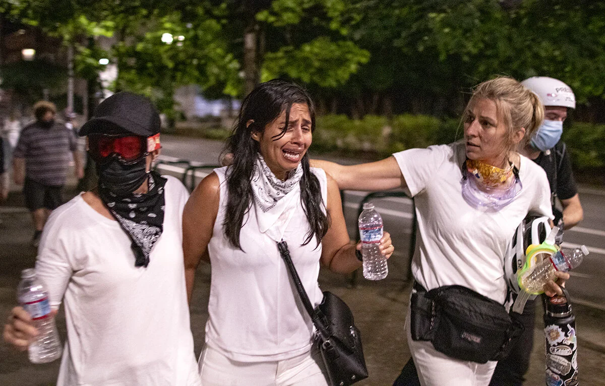  Juli Maus (left) and Erin Kelley (right) help a protester (center, name withheld) who was blinded by tear gas fired by federal police officers. Roughly 500 people gathered Sunday, July 18, on the 52nd consecutive night of protests at the Multnomah C
