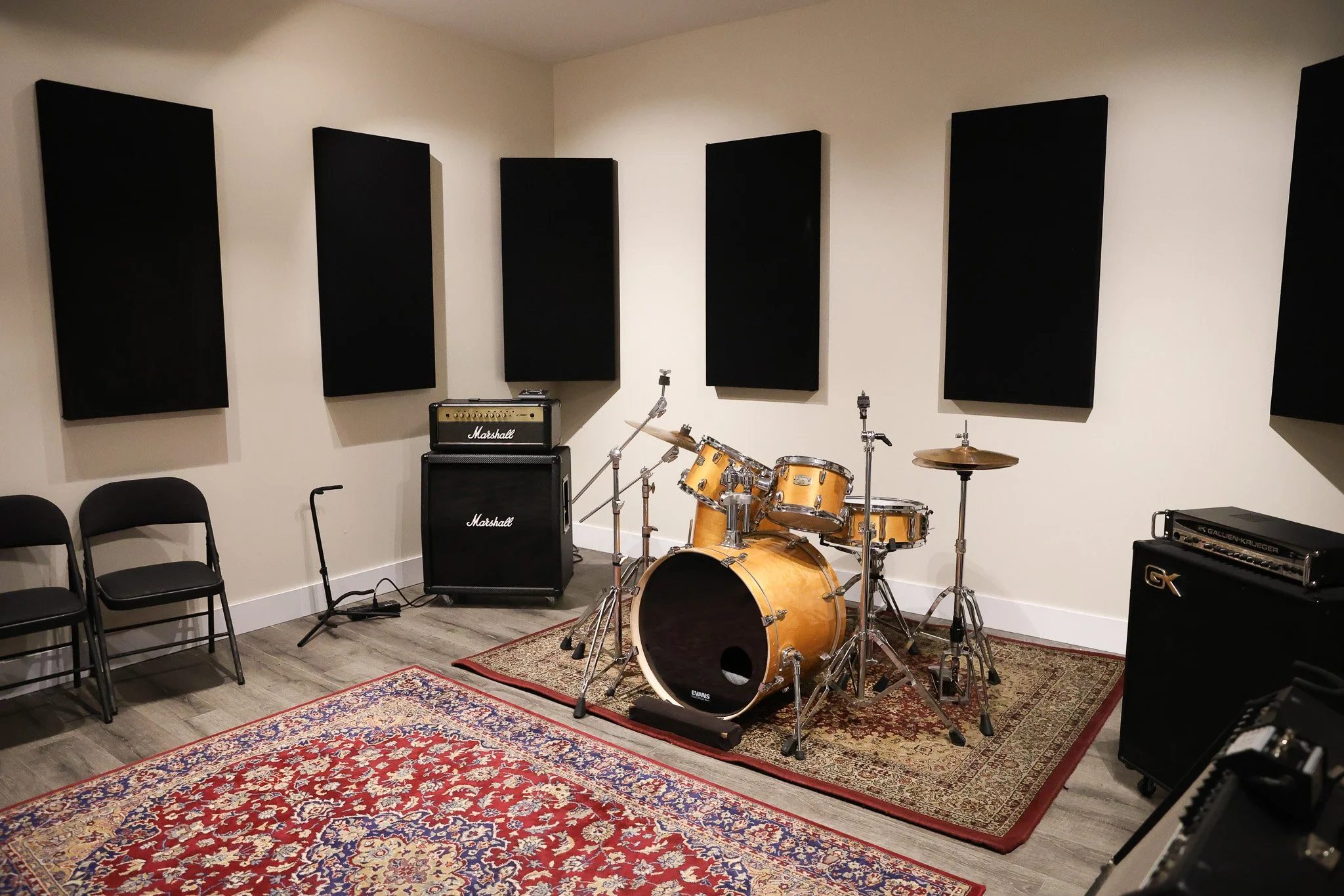 Music practice room with musical instruments, amplifiers, and black soundproof panels on the white wall, and a colorful oriental rug on the floor.