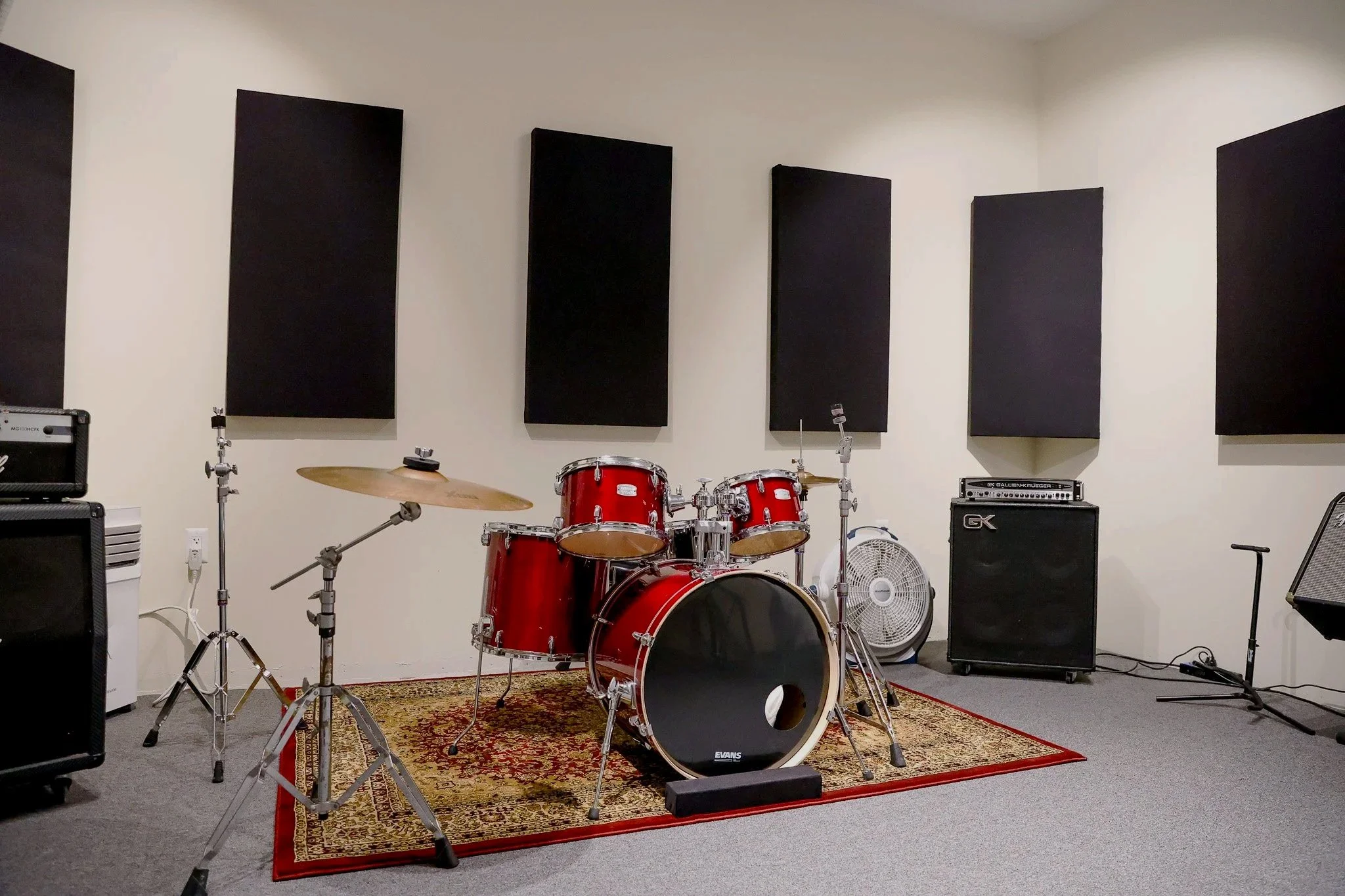 Music rehearsal room with a red drum set, black amplifiers, a microphone in front, a guitar amplifier, music stands, and acoustic panels on the white walls.