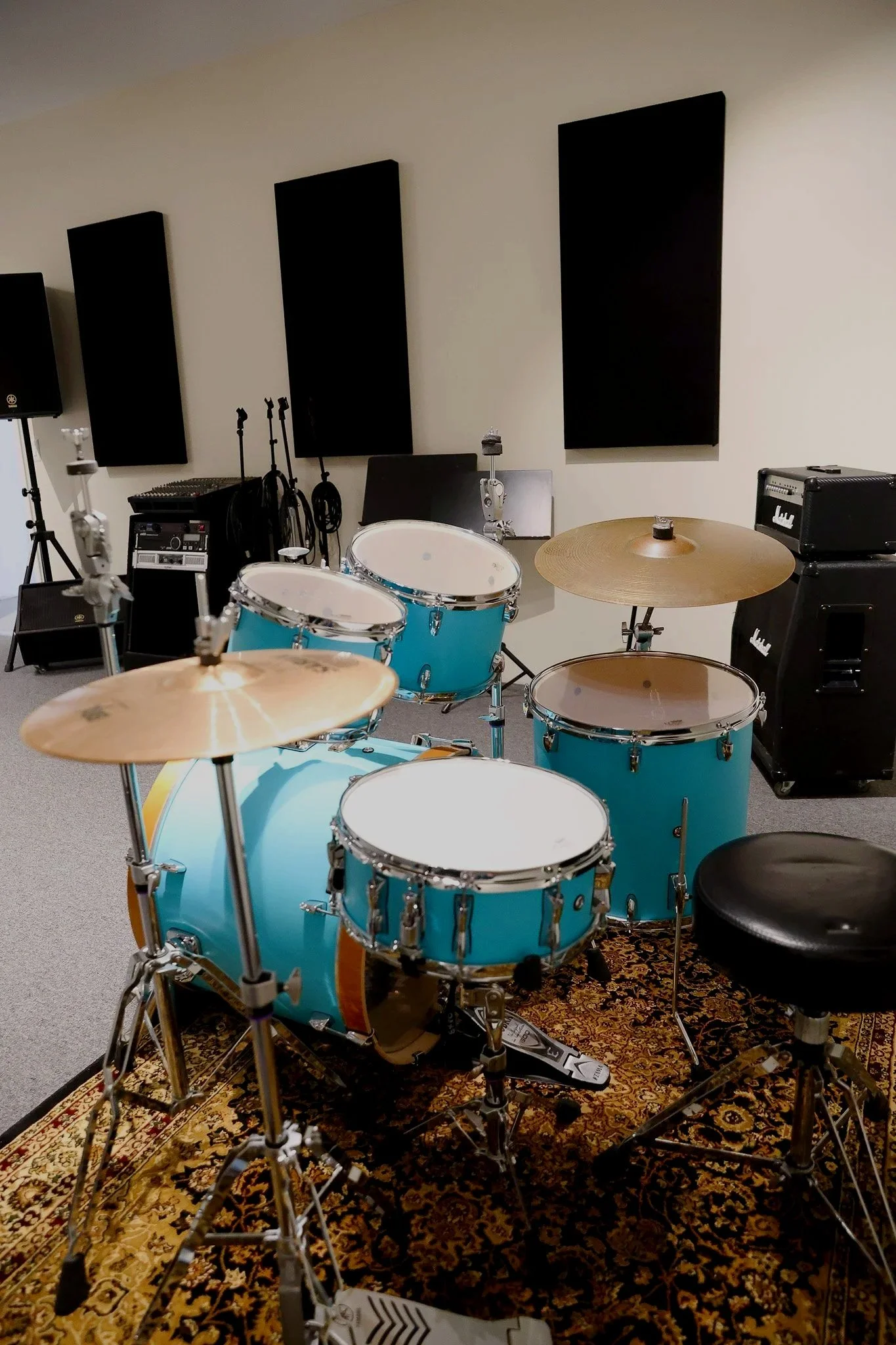 A music practice room with a blue drum set on a patterned rug, black soundproof panels on the wall, and various musical equipment including speakers, microphones, and amplifiers.