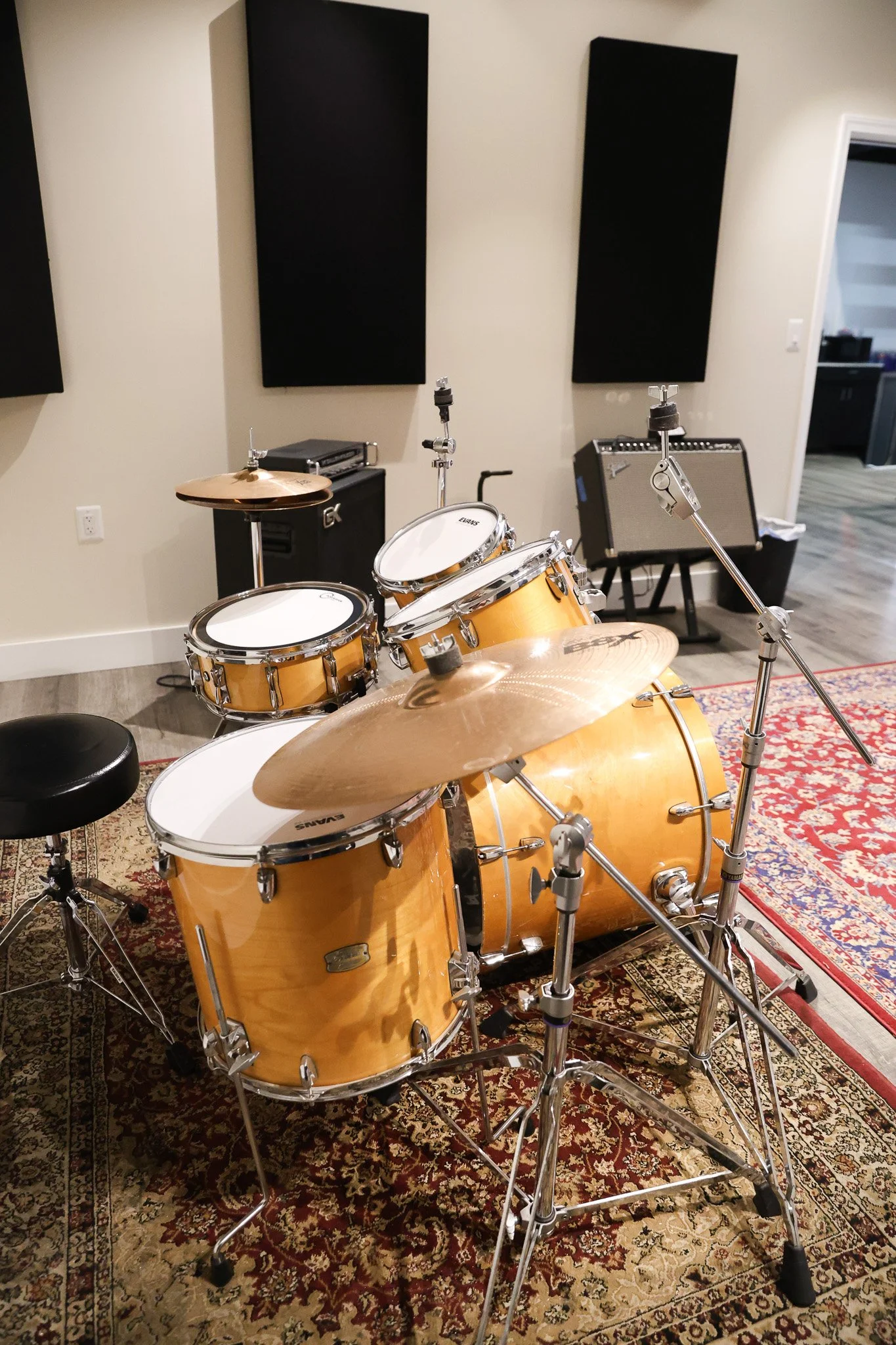 A music rehearsal room with a drum set, guitar amplifier, and acoustic panels on the walls. The drum set is golden brown with multiple drums and cymbals, placed on a patterned rug.