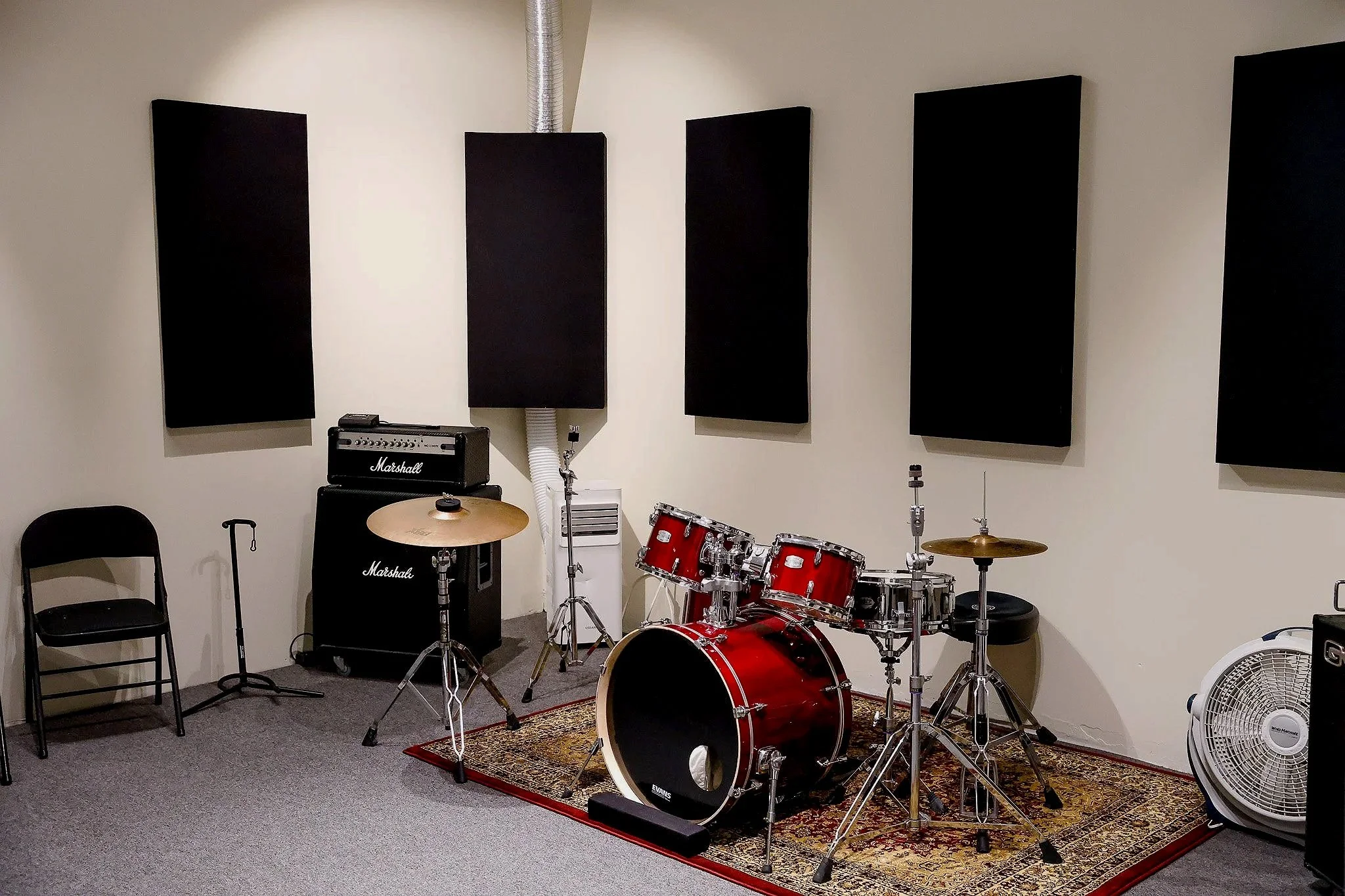 A music practice room with a red drum set on a patterned rug, a Marshall amplifier, black acoustic panels on the wall, and various musical equipment.
