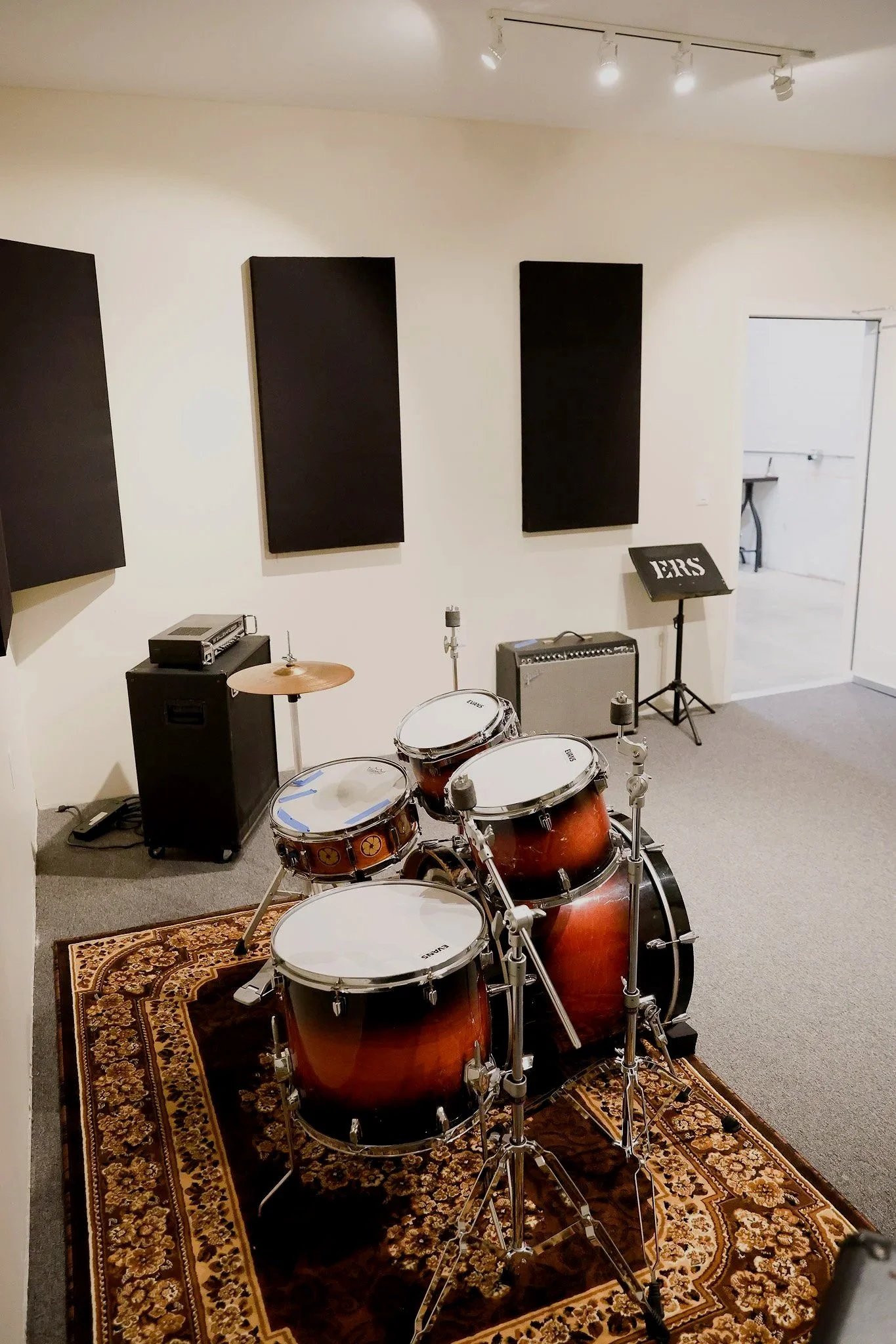 A music rehearsal room with black acoustic panels on white walls, a rug with intricate patterns on the carpet, and a drum set placed on the rug. There is a music stand with the word 'PERS' on it, and variety of musical equipment including a guitar amplifier and a cymbal. The room appears to be used for band practice.