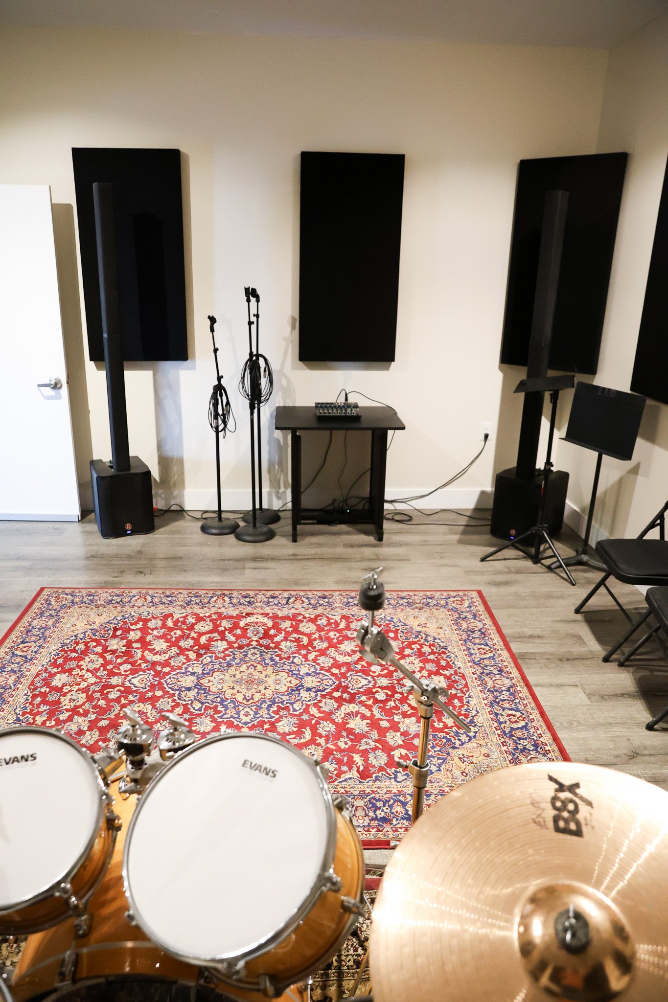 Band room with black acoustic panels, microphones, drum set, and piano, viewed from behind the drum kit, with a red patterned rug on the floor.