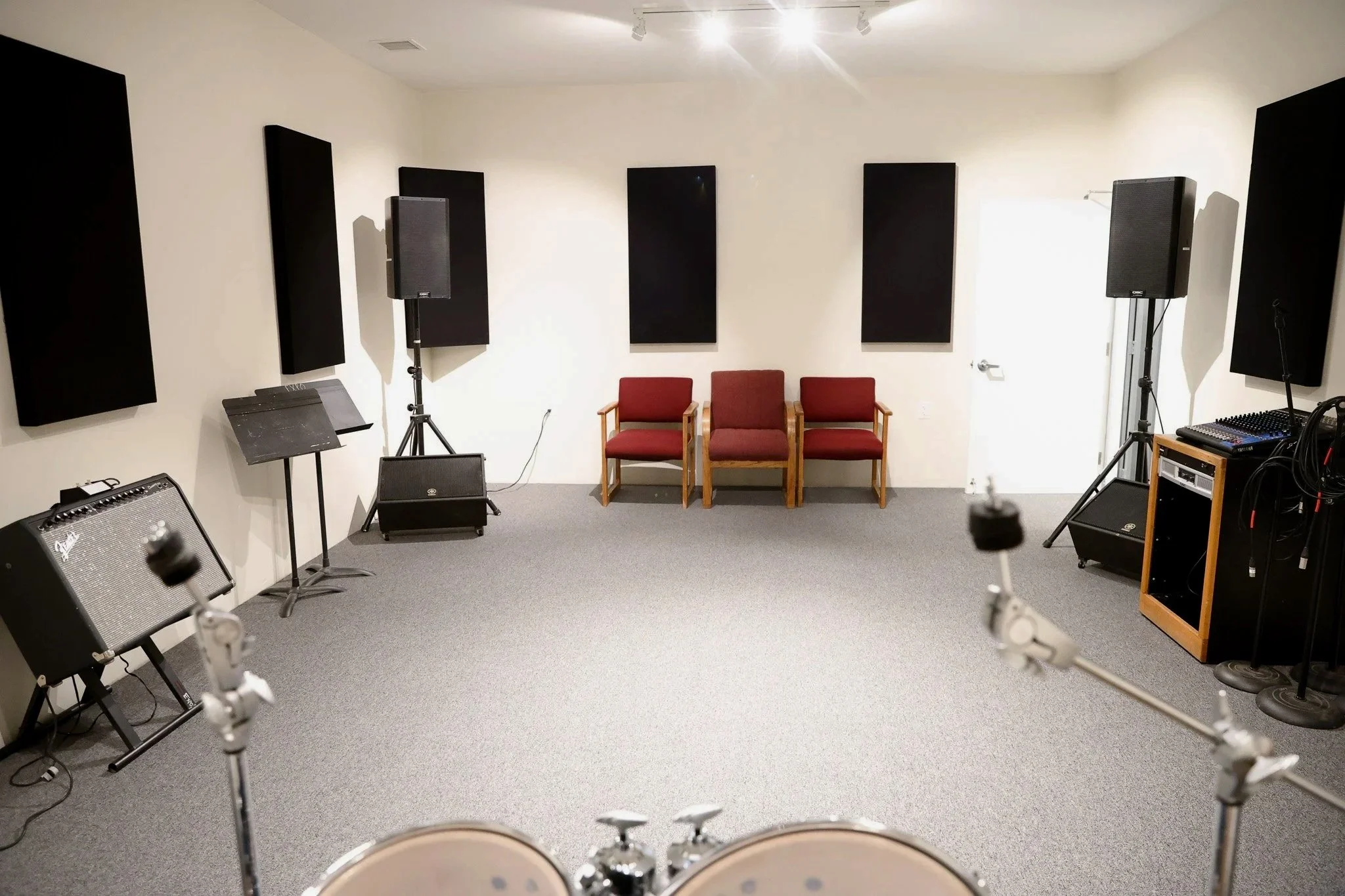 Music rehearsal room with wall-mounted black sound panels, a drum set in the foreground, chairs against the wall, microphones, amplifiers, and music equipment.