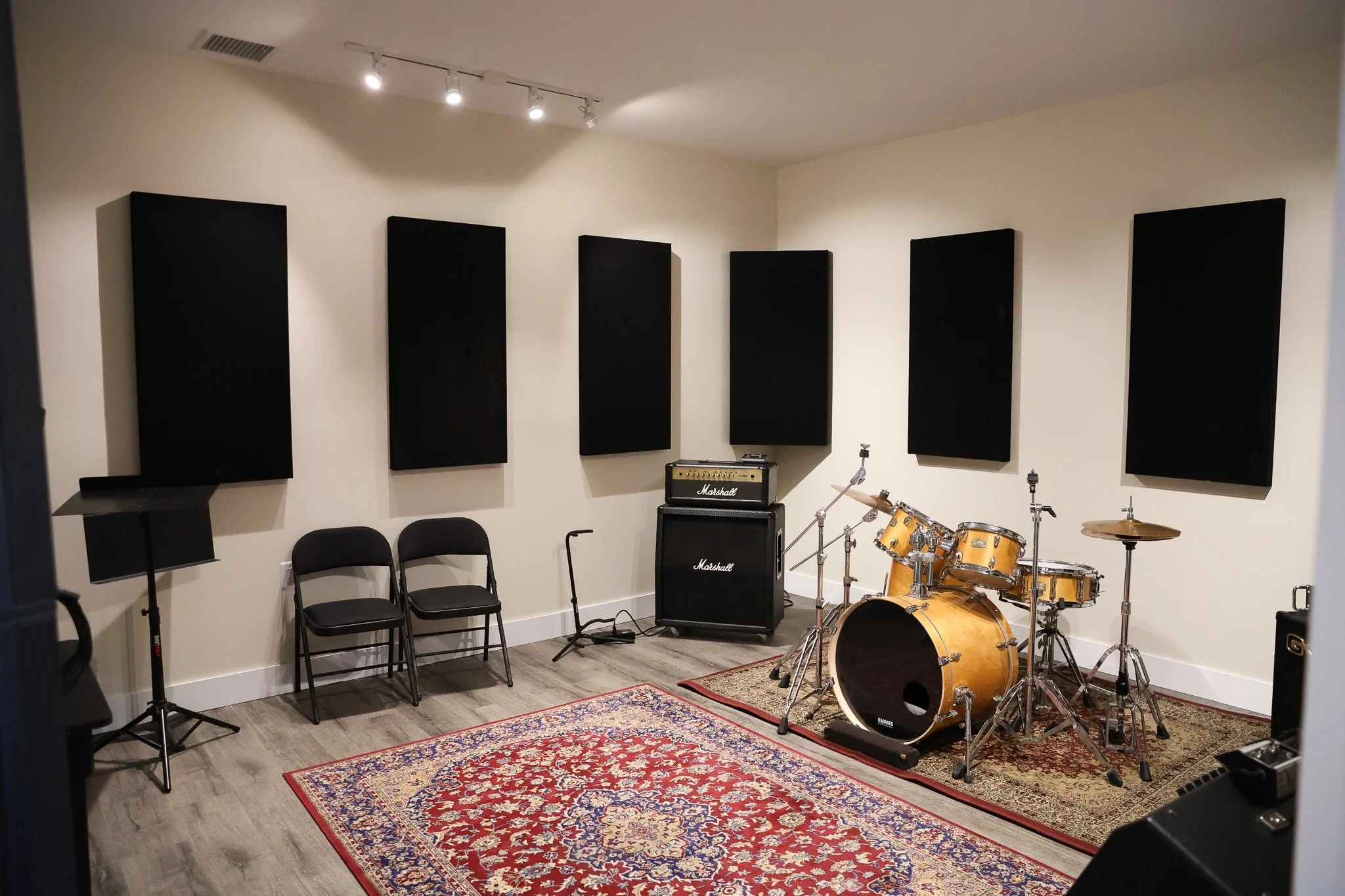 Empty music practice room with black acoustic panels on white walls, musical instruments including a drum set, guitar amplifiers, and chairs, with a red patterned rug on the wooden floor.