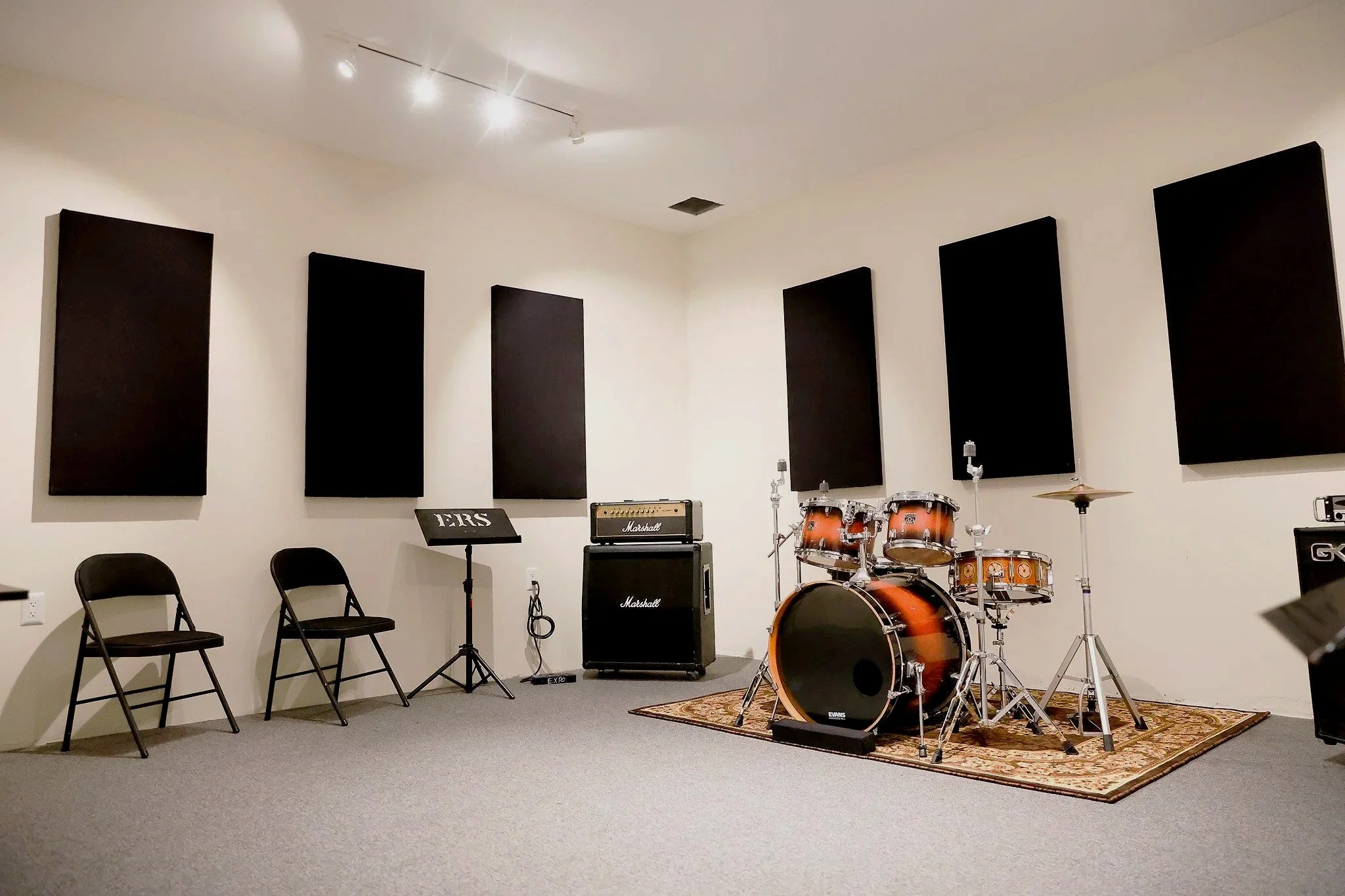An indoor music rehearsal space with black acoustic panels on the white walls, chairs lined up on the left, and musical equipment including drums, amplifiers, and a music stand on a rug on the right.