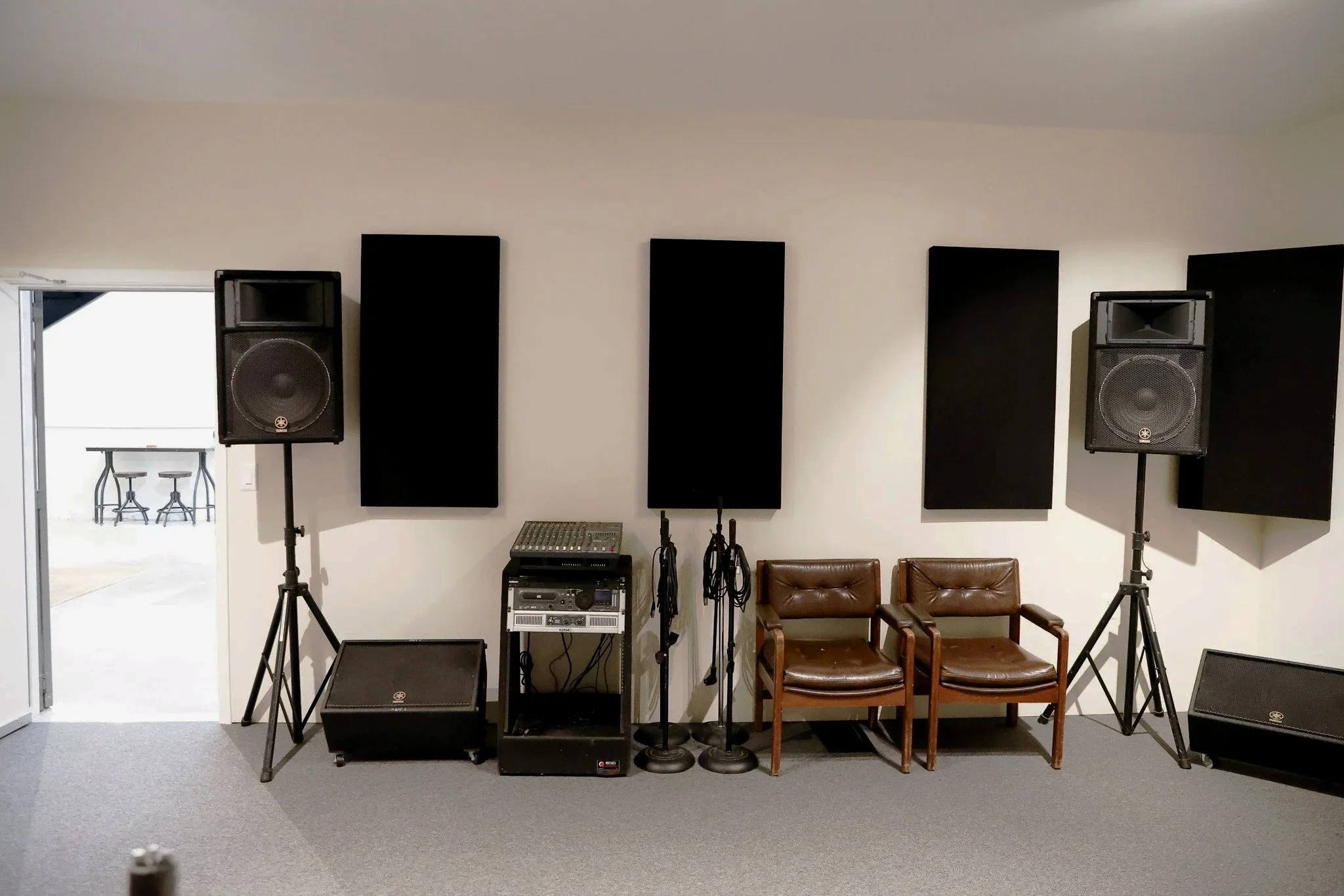 A music rehearsal room with two large black speakers on stands, black acoustic panels on the wall, a small mixer and audio equipment, two wooden chairs, and a doorway leading to another room with two stools and a table.