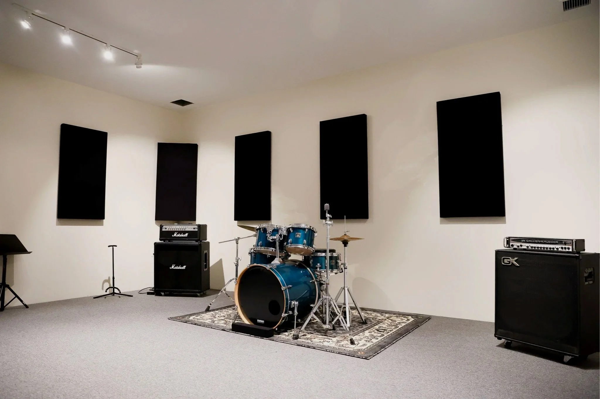 A music practice room with black acoustic panels on a white wall, a drum set on a decorative rug, and multiple amplifiers and musical equipment.