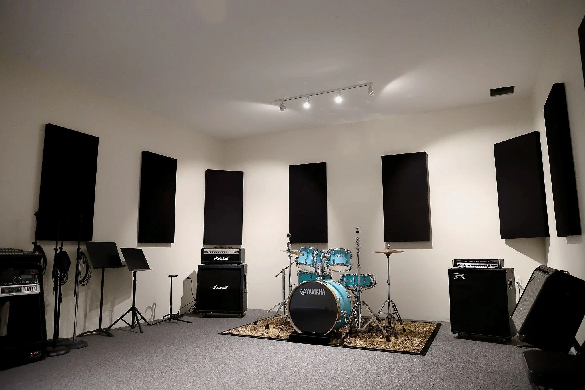 An empty music rehearsal or recording room with musical instruments and equipment. There are black acoustic panels on the white walls, a drum set in the center, guitar amplifiers, microphones, and music stands. Overhead lights illuminate the room.
