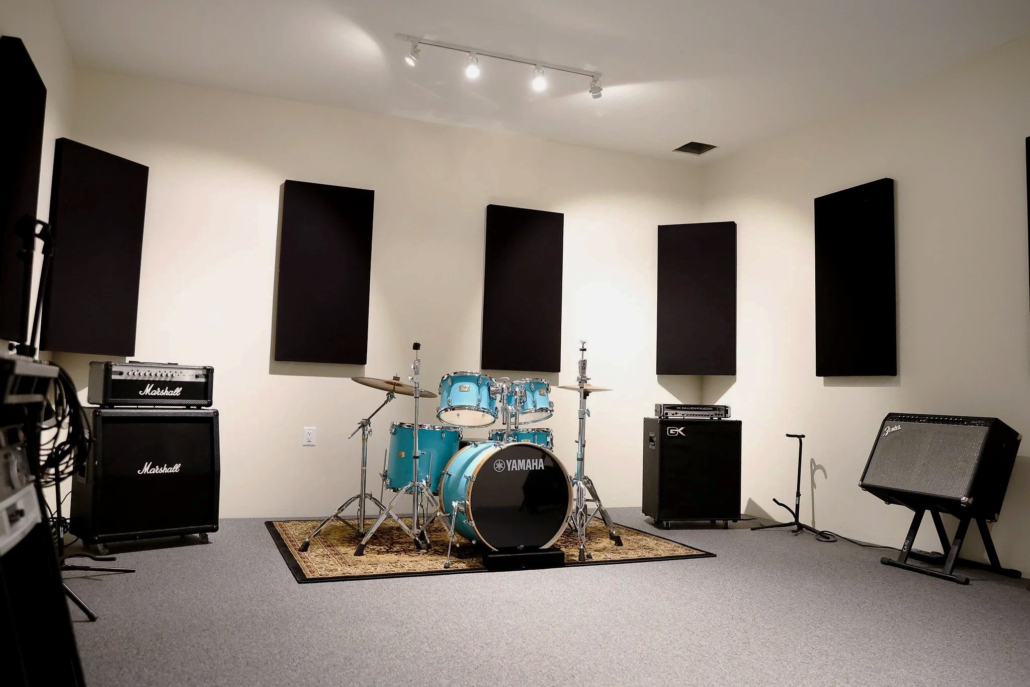 A music practice room with a Yamaha drum set in the center, surrounded by Marshall and GK guitar amplifiers, black acoustic panels on the walls, a keyboard on a stand, and various cables and equipment in the room.