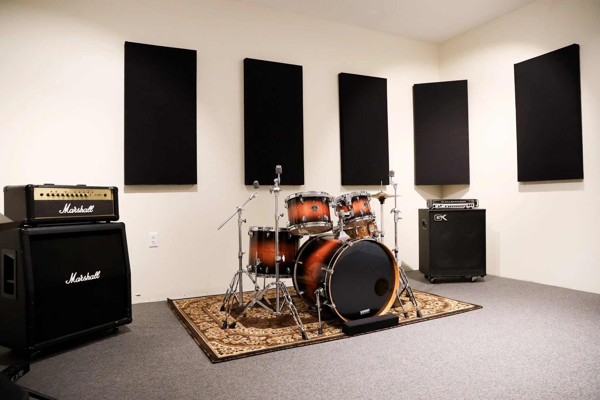 A music rehearsal room with black soundproofing panels on the white wall, a drum set on a patterned rug, and Marshall amplifiers.