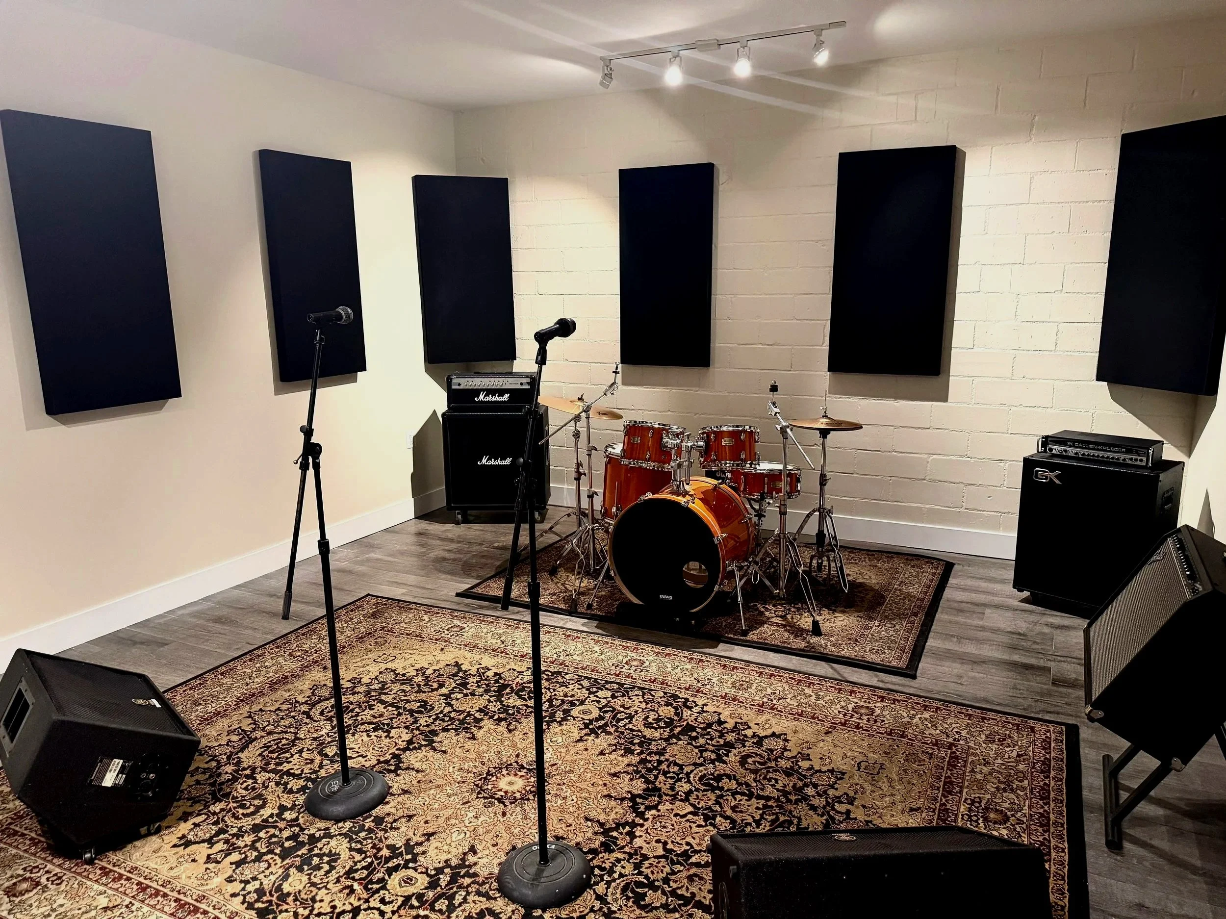 A music rehearsal room with a drum set, speakers, microphones, and acoustic foam panels on the wall.