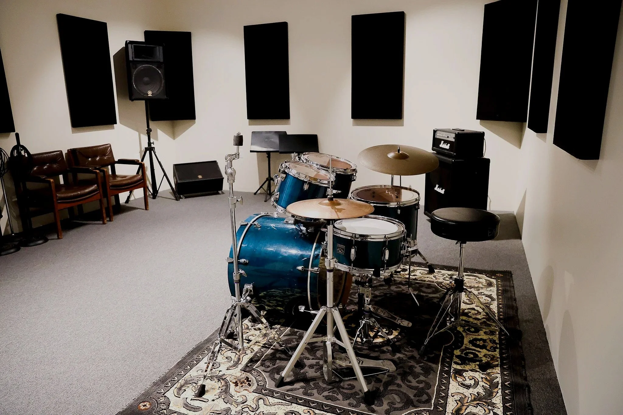 Music rehearsal or recording studio with a drum set in the foreground, two brown chairs, black soundproof wall panels, and audio equipment.