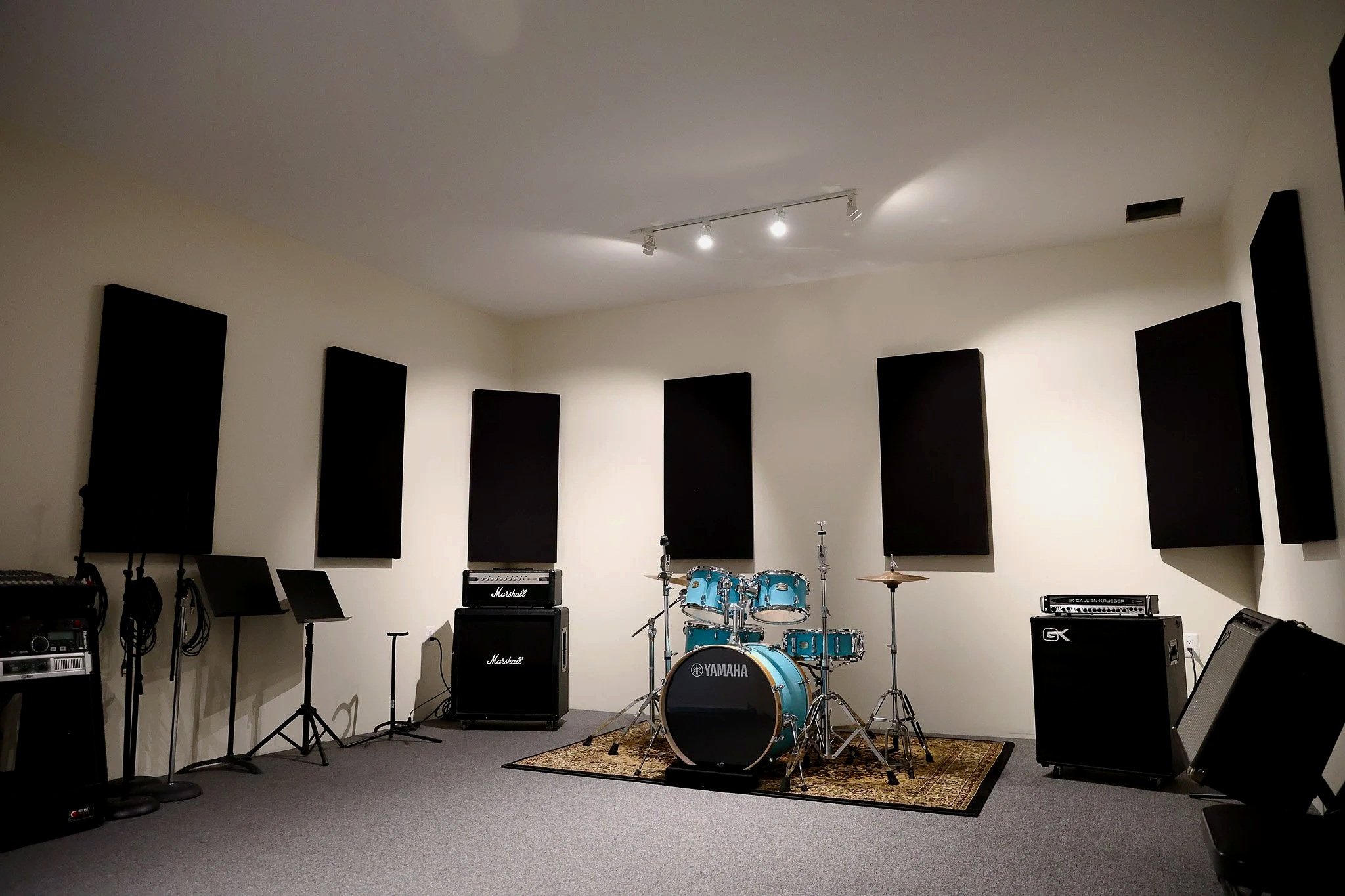 A music rehearsal room with black acoustic panels on the white walls, musical instruments including a drum set, guitar amps, and microphones, and music stands. The room has carpeted flooring and is illuminated by ceiling track lights.