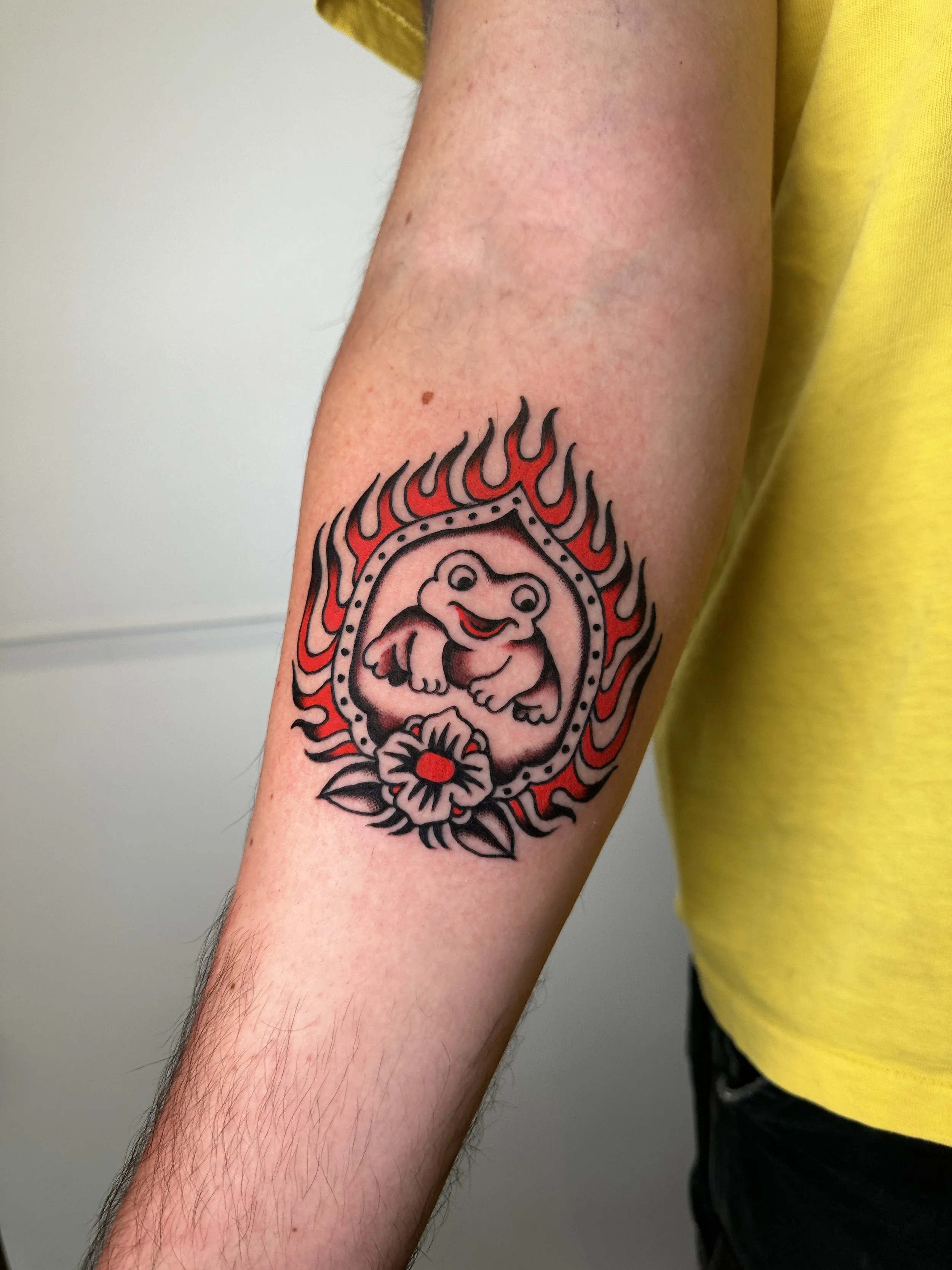 Tattoo of frog framed by flames and a flower on lower arm.  