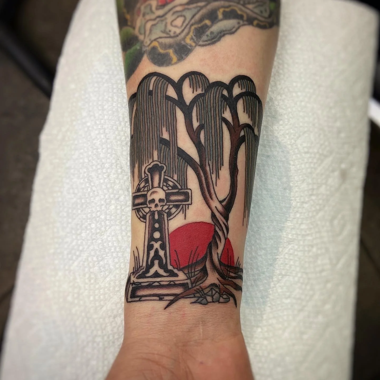 Tattoo of celtic cross statue with skull in the center next to a tree with a red sun setting in the background