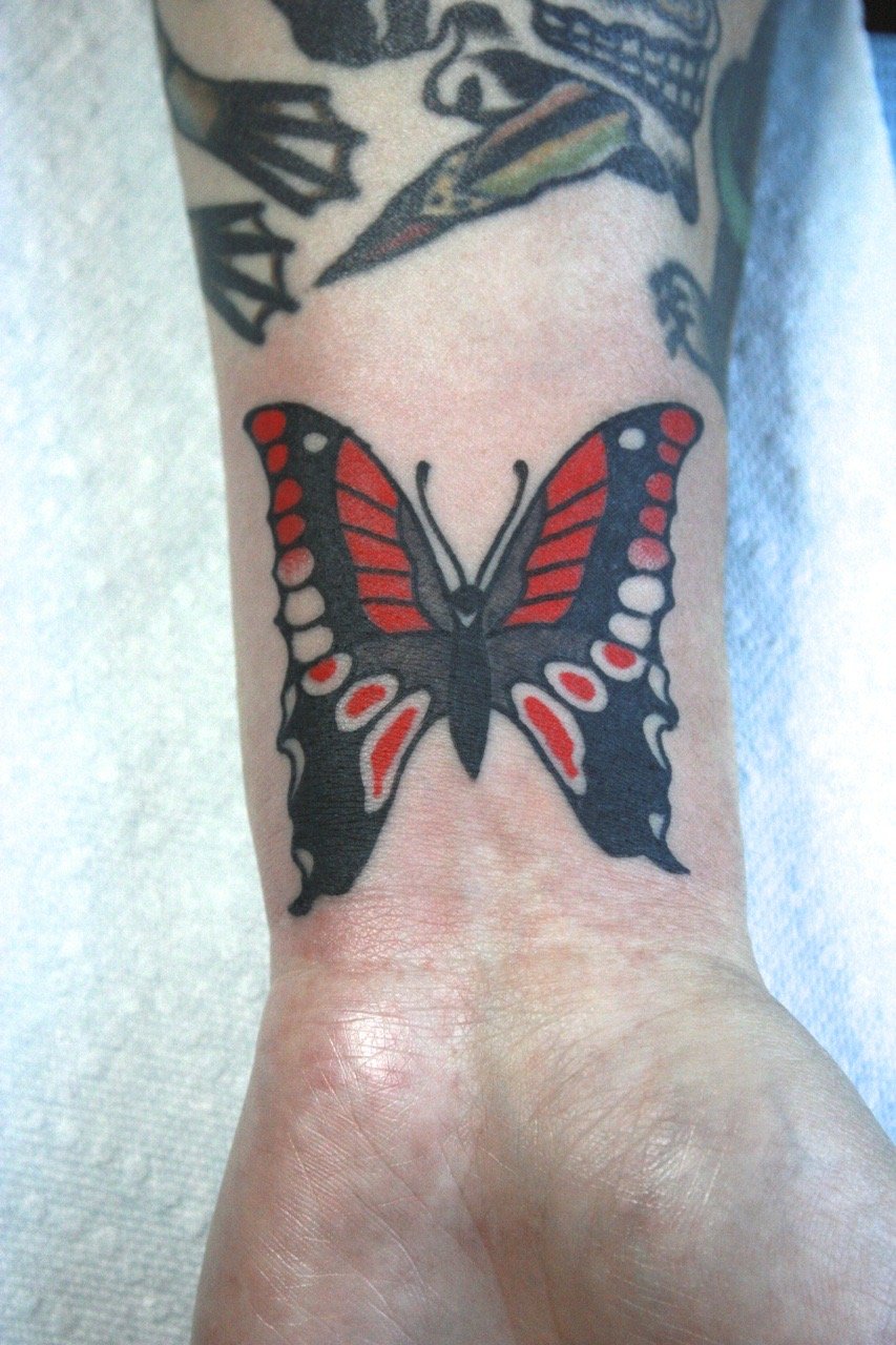 Tattoo of butterfly in black, grey, and red on the wrist. 