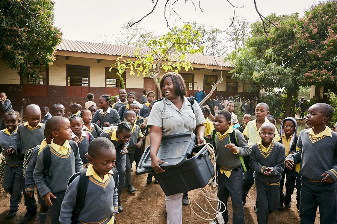 Students at Phalaborwa Primary School crowd around Bush Babies education officer Lewyn Maefala who carries a plastic box filled with elephant dung, South Africa, 2017. In the background, Herman, a Norwegian volunteer, carries Lewyn’s projection screen. Most days, however, she is on her own.