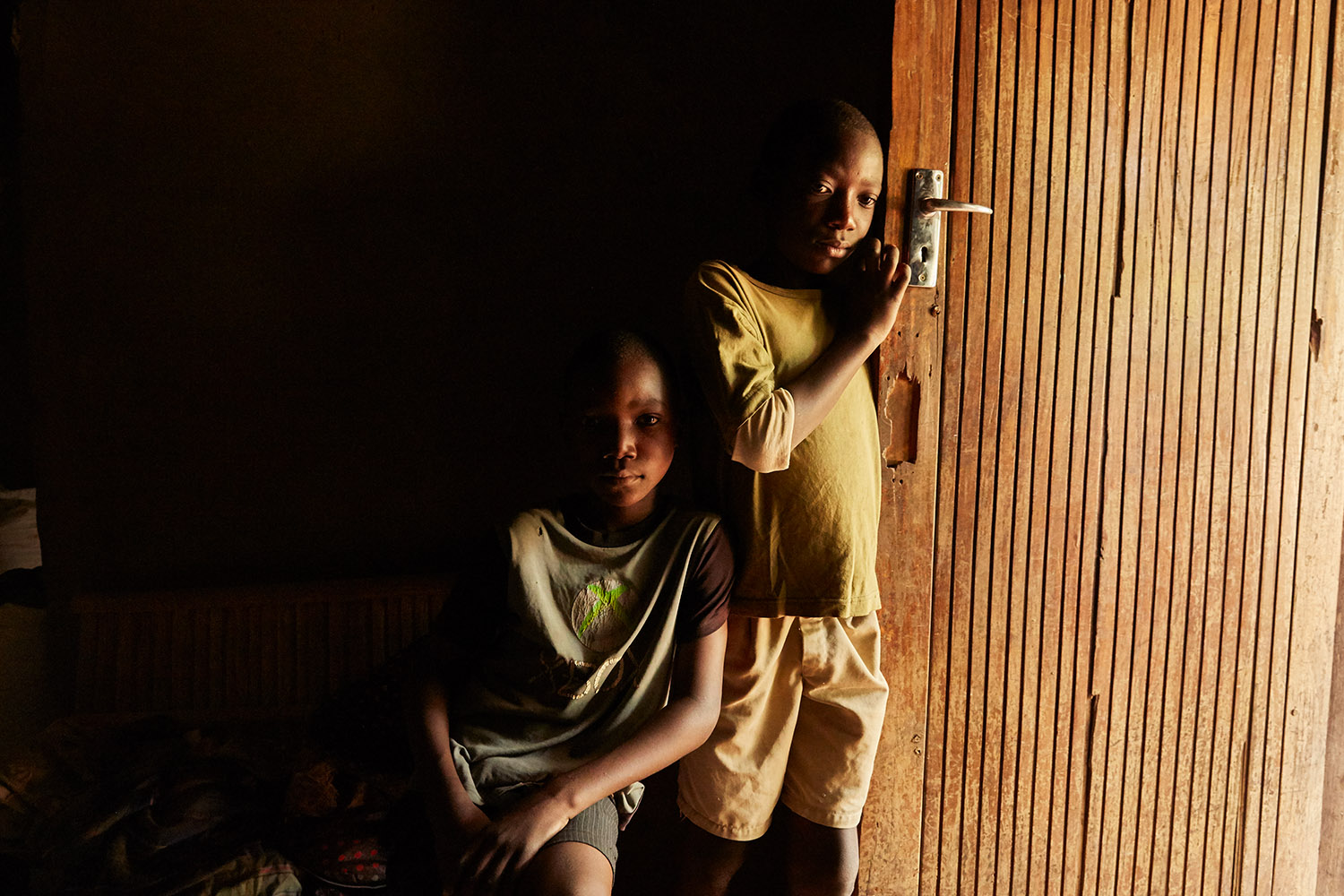  Jan Chumba (12) and his brother Ben (10) in the doorway of their home, Chiuta village, Malawi, 2017.  During the day, the only light inside comes from the open door and windows. 