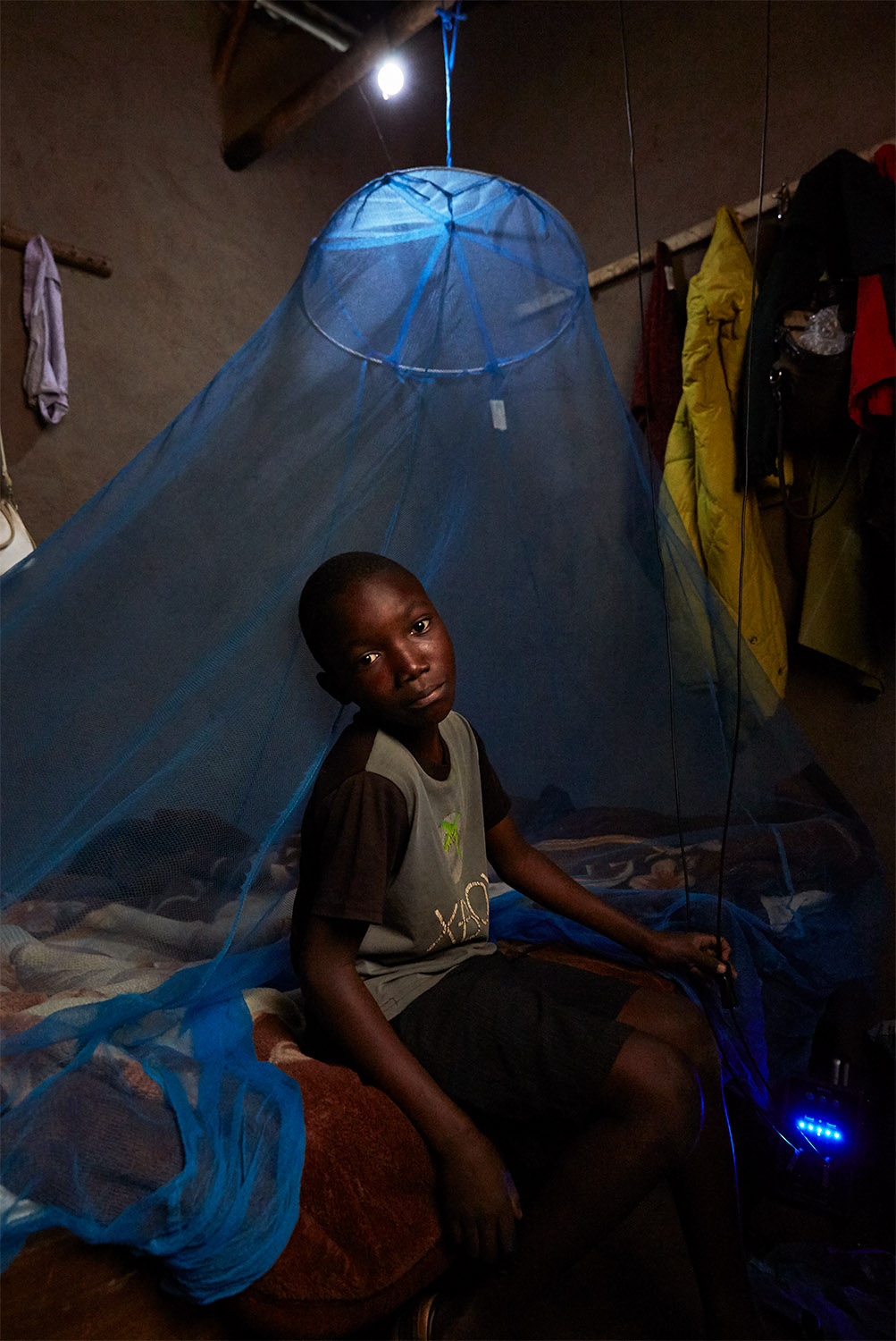  Ben Chumba (10) sits on his bed, lit by a light bulb which is powered by a solar battery, visible in the bottom right hand corner of the image, Chuita village,  Malawi, 2017. 