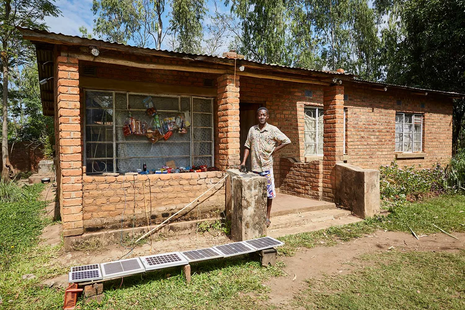 Felix Wonga (29) stands next to the small store he runs with his father, Robert (56), Luchenza, southern Malawi, 2017. Felix runs the mobile phone charging business, while his father takes care of the shop front. The solar panels laid out in front of the store power a car battery which, Felix uses to charge up to five mobile phones at once. Because Felix and Robert’s home is the only one in the area with solar panels, business is good.