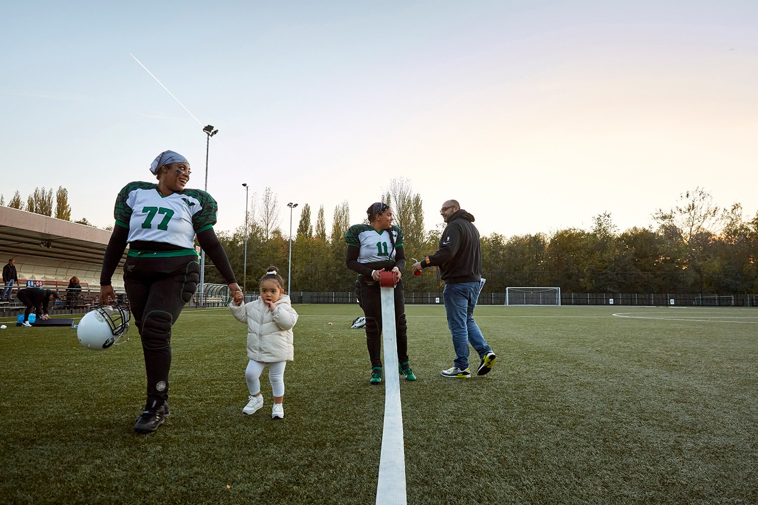  Marjaline, center for the Rotterdam Ravens, walks off the field with the daughter of a friend after a game against the Utrecht Wolverines, Rotterdam, NL, 2021. Behind her, Lisa, one of the Ravens' tight ends, chats with the game's live commentator w