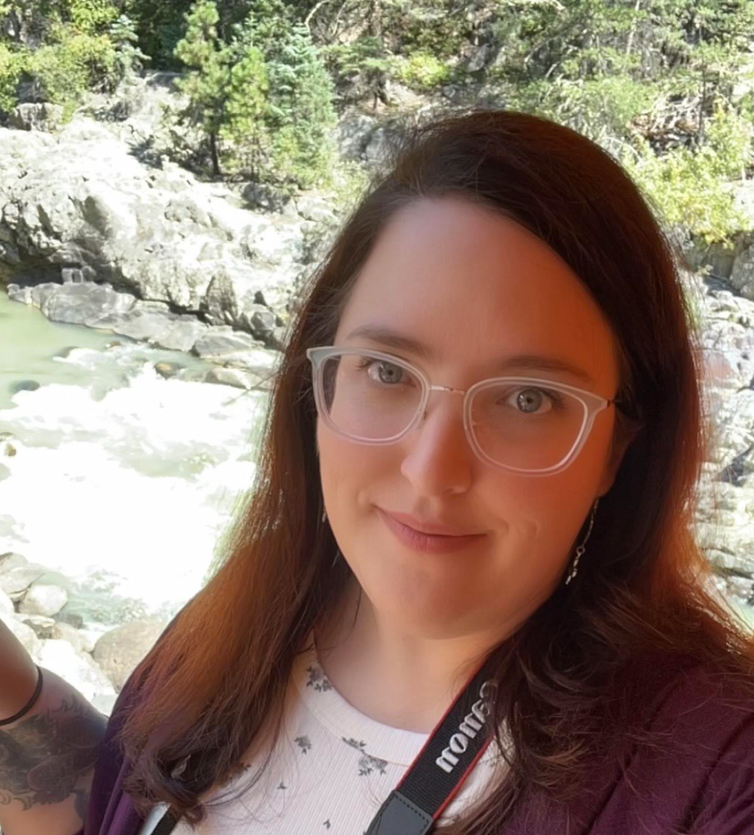 A woman wearing glasses and a white shirt with small floral patterns, smiling outdoors with a river and rocks in the background.