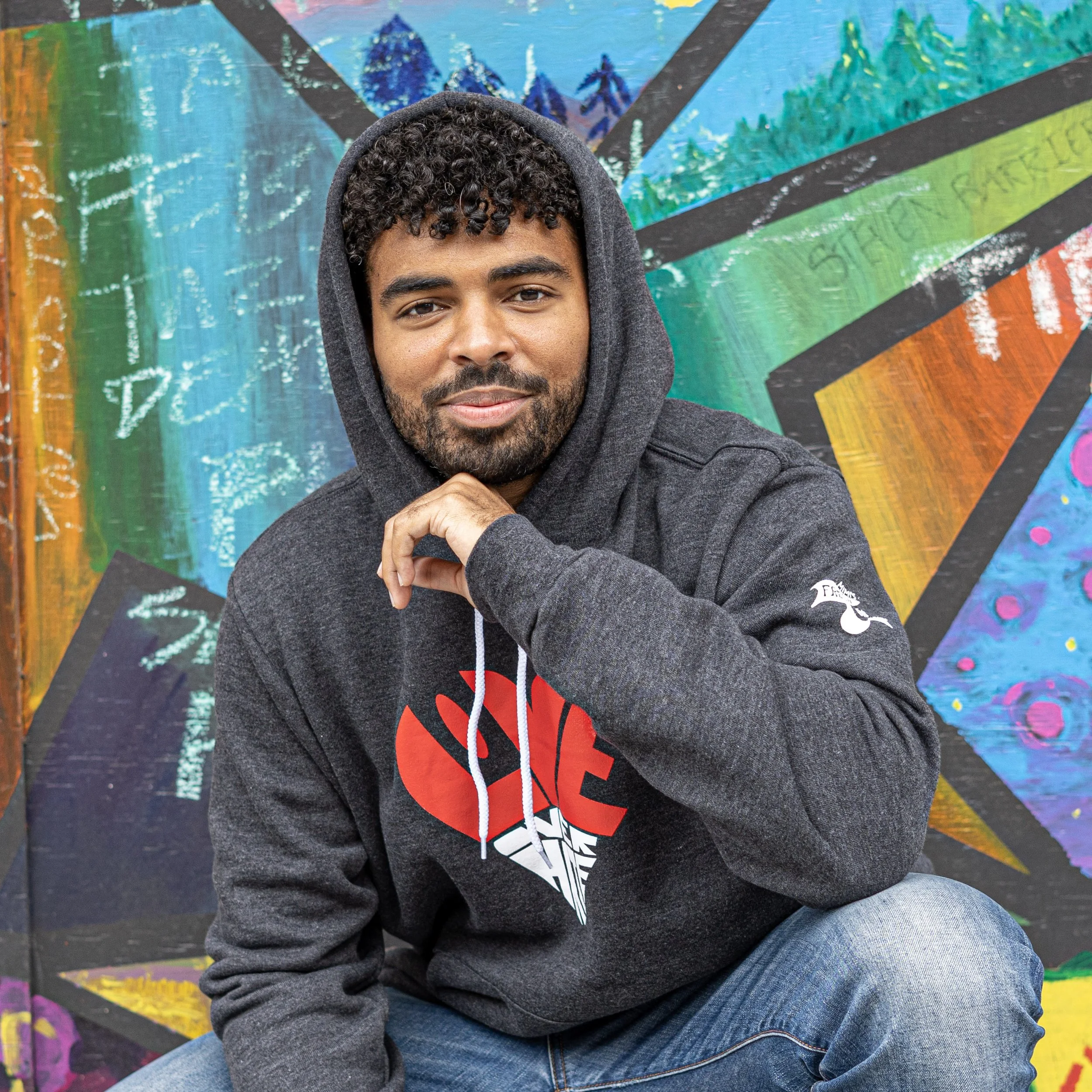 A young man with curly hair and a beard sitting in front of a colorful, abstract painted mural. He is wearing a dark hoodie and jeans, resting his chin on his hand, and looking at the camera.