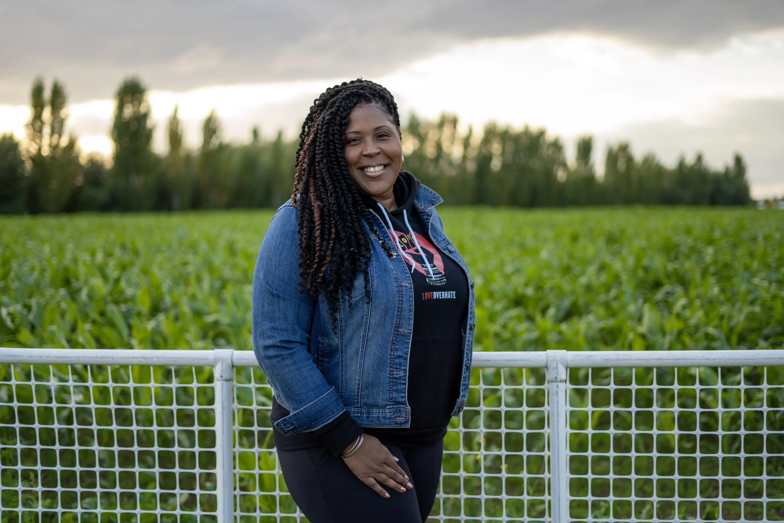Smiling woman with braided hair standing outdoors in front of a green field and trees with a cloudy sky.
