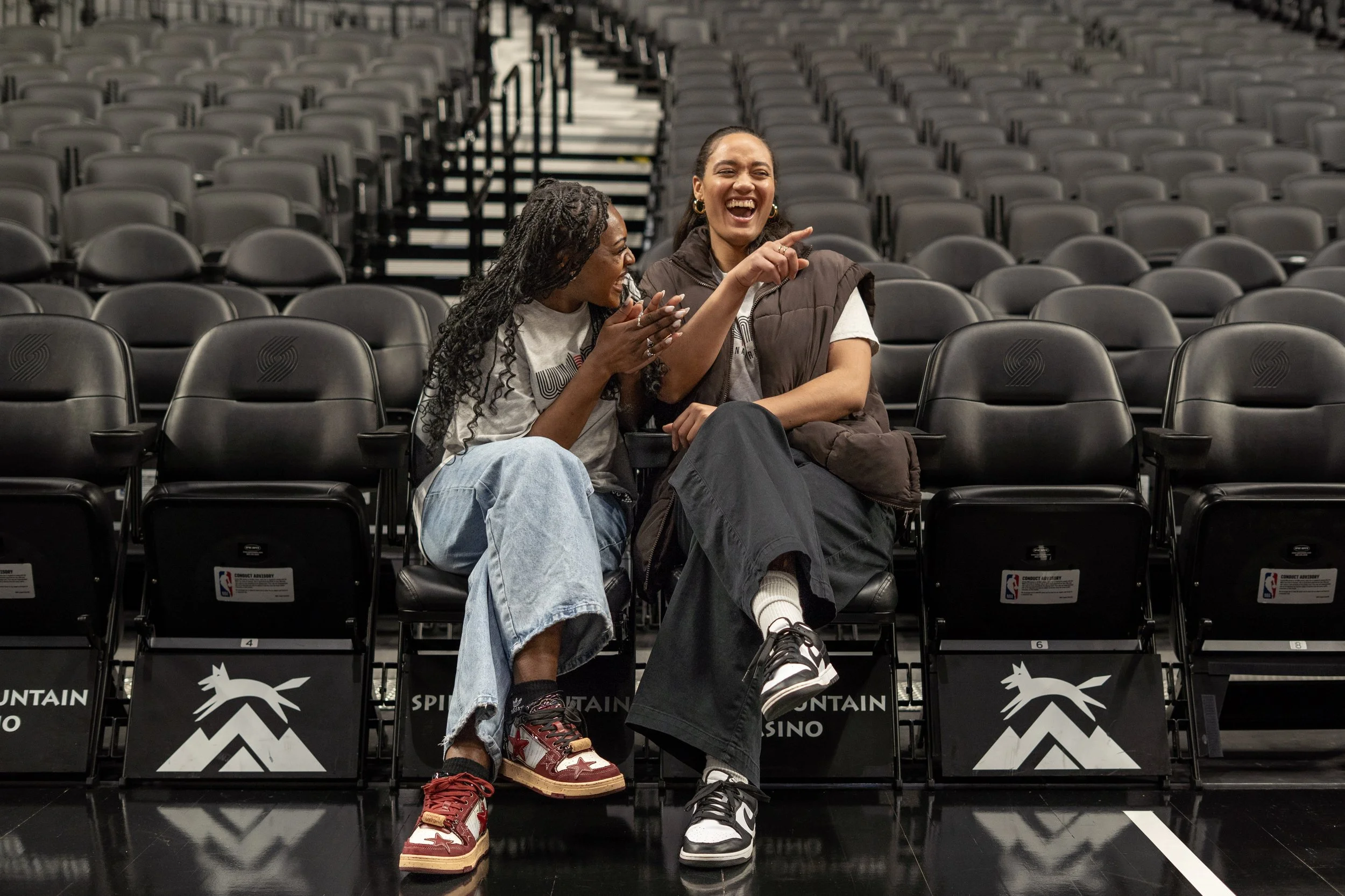 Two women laughing and talking while sitting in empty stadium seats, with one pointing at the other.