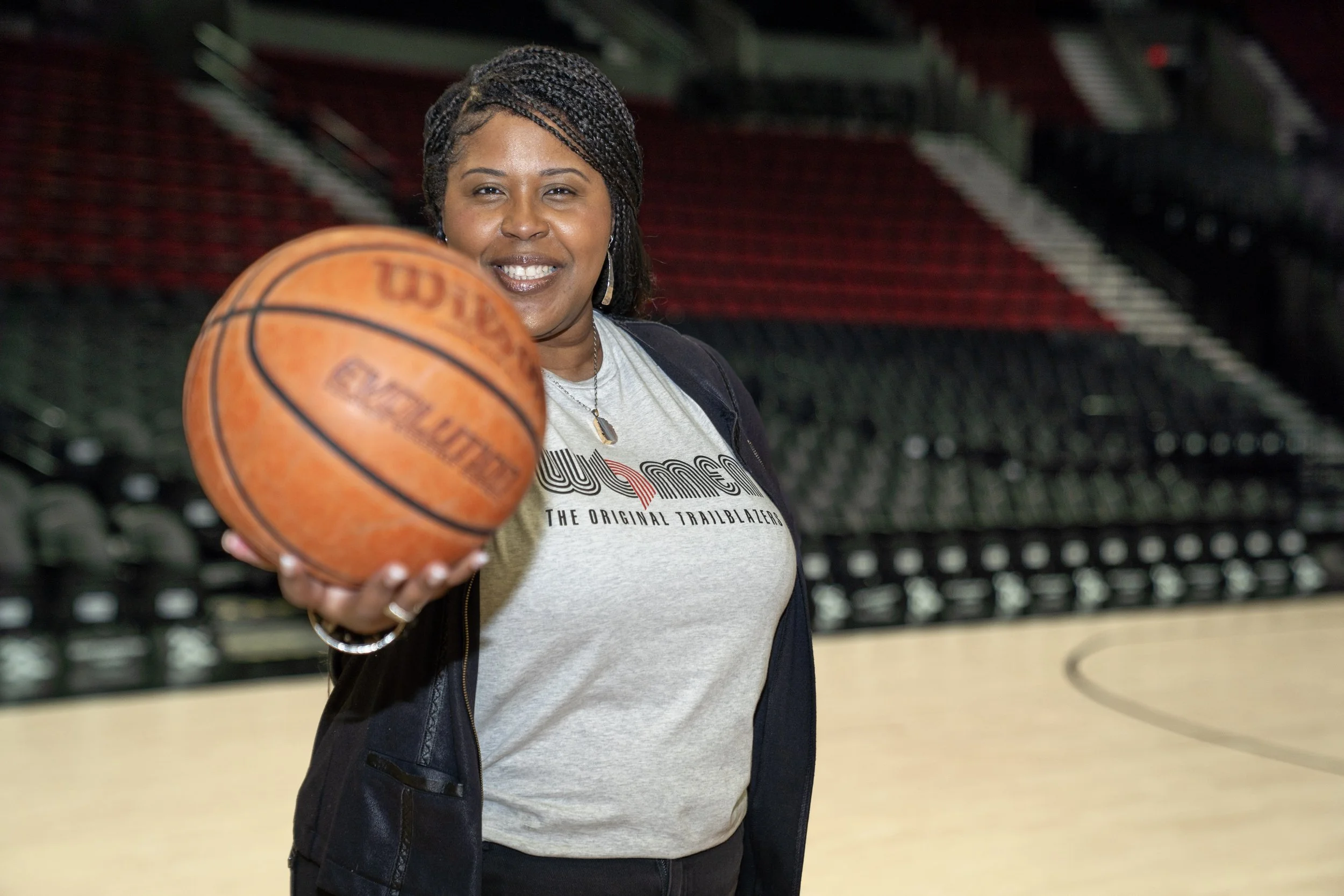 A woman with braided hair smiling and holding a basketball in a sports arena.