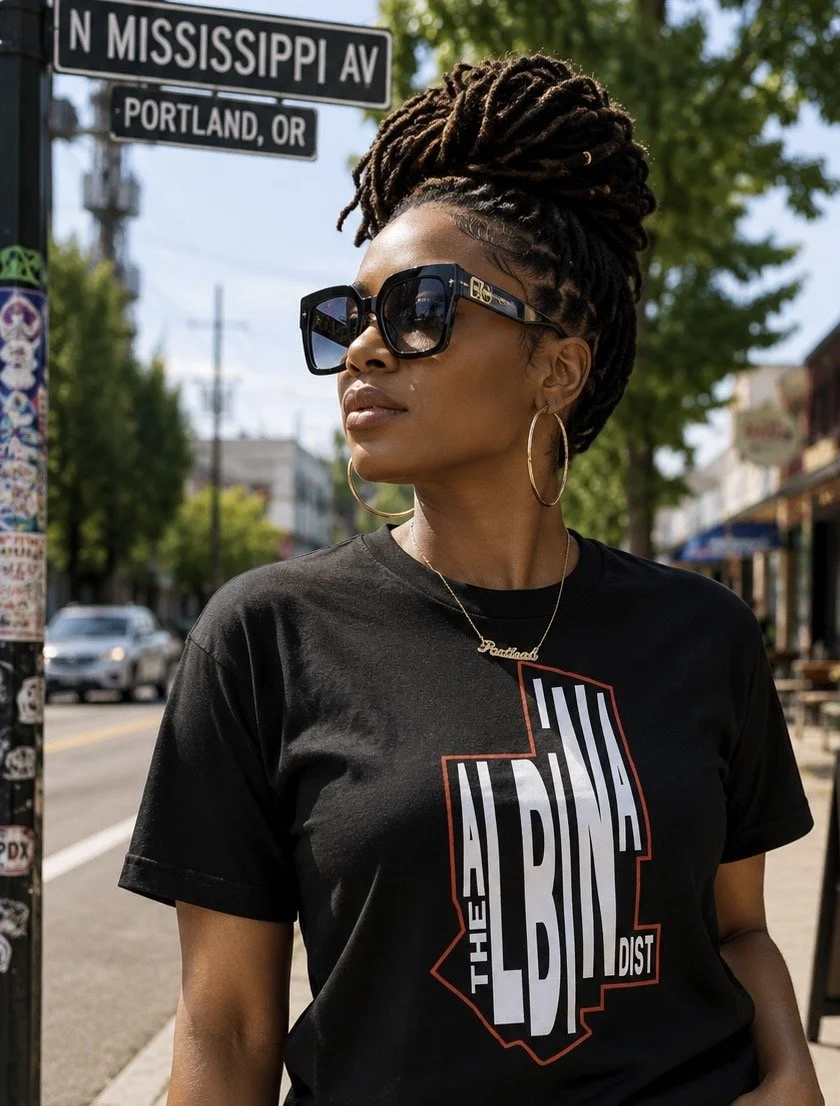 A woman with dreadlocks styled in a high bun wearing large sunglasses, hoop earrings, a gold necklace with a name tag, and a black t-shirt with the words 'The Land of the LDNA' printed on it, standing outdoors on a city street in Portland, Oregon.