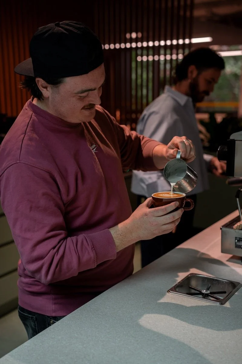Barista practising a milk pour at a café coffee station while another person works nearby.