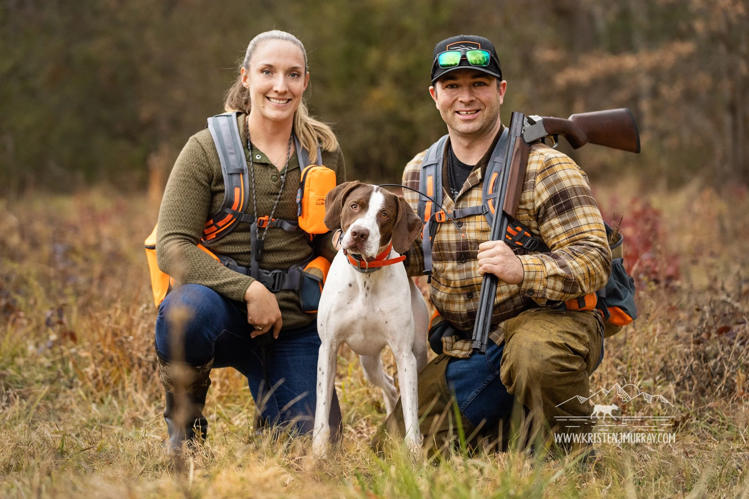 A man and woman with a hunting dog posing in a forested area during fall, both wearing outdoor gear and backpacks, with the man holding a rifle. Upland Hunting in Virginia