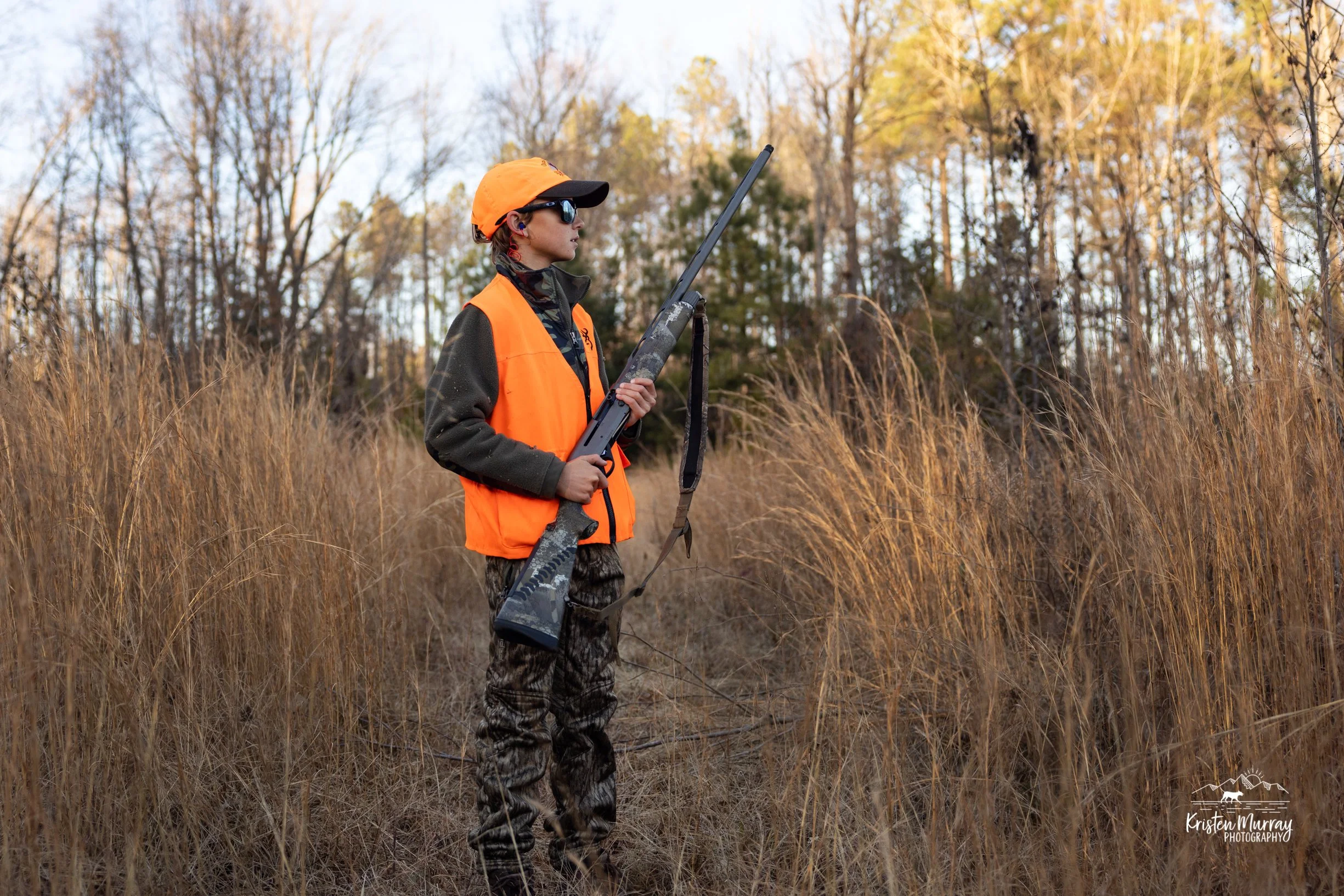 A person in camouflage pants, orange vest, and orange cap holding a hunting rifle, standing in tall grasses in a forested area during autumn.