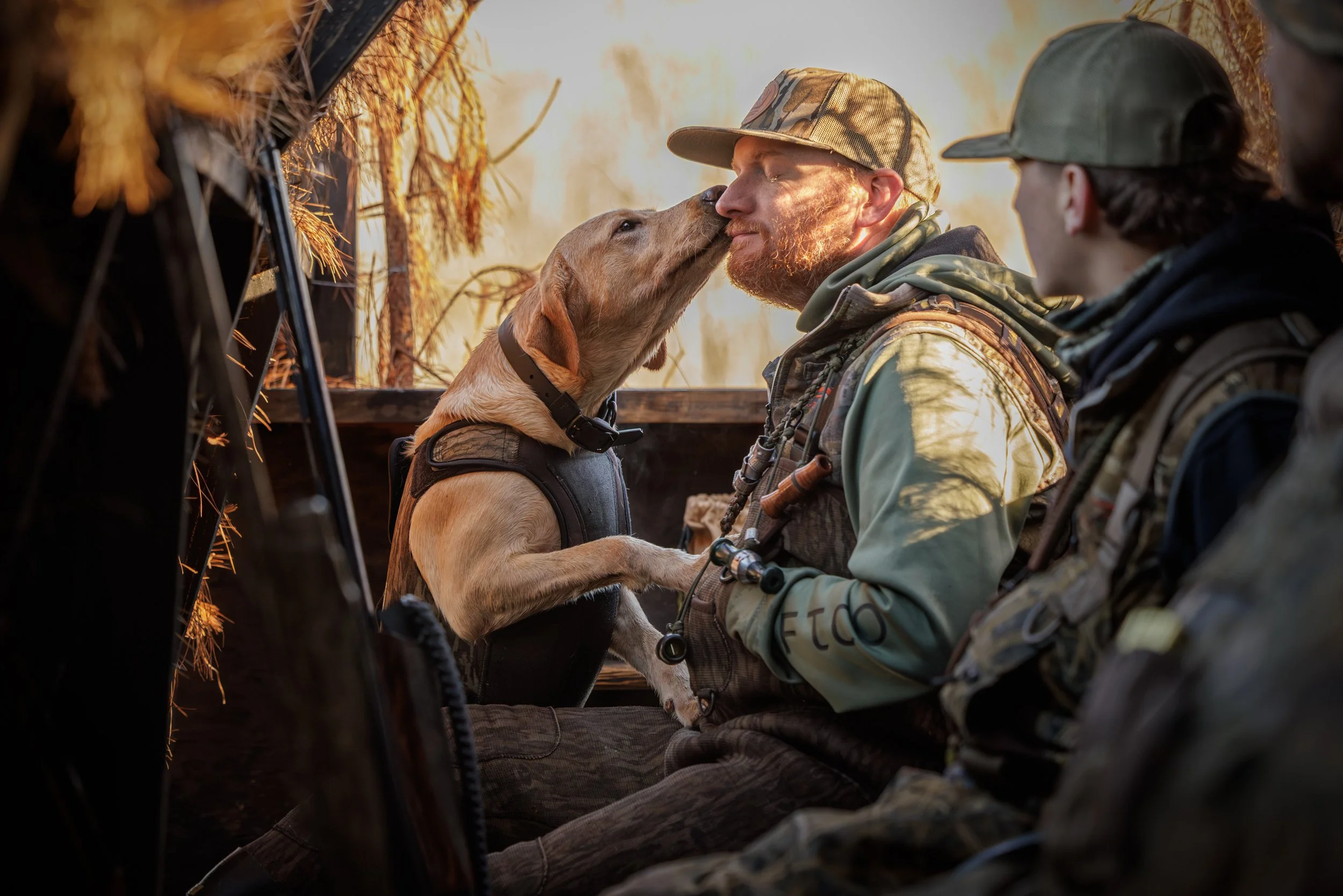 A man with a beard and camouflage cap kissing a yellow Labrador retriever in a hunting blind with two other people nearby, during sunset.