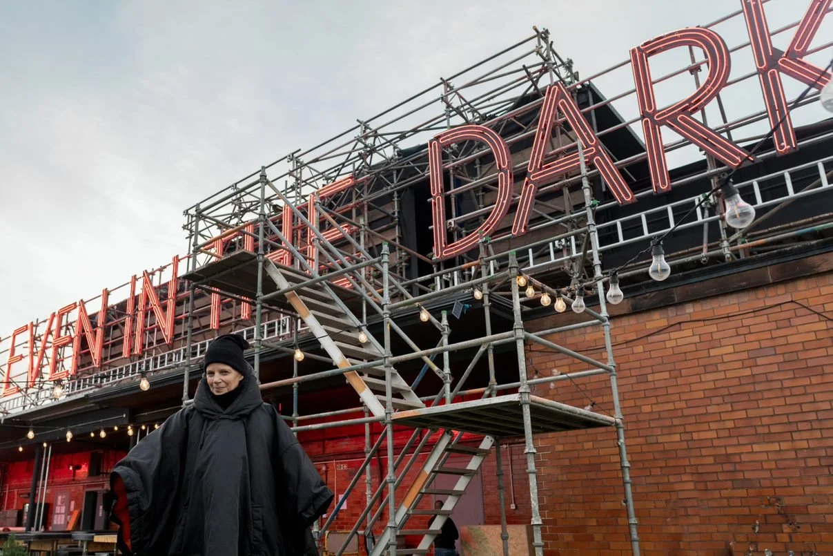 Mary Ellen Carroll standing in front of large bright neon letters spelling out the phrase 'even in the dark'