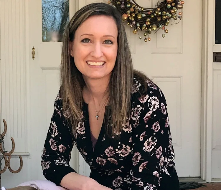 A smiling woman with shoulder-length brown hair wearing a floral black cardigan, sitting indoors near a door with a decorative wreath on it.
