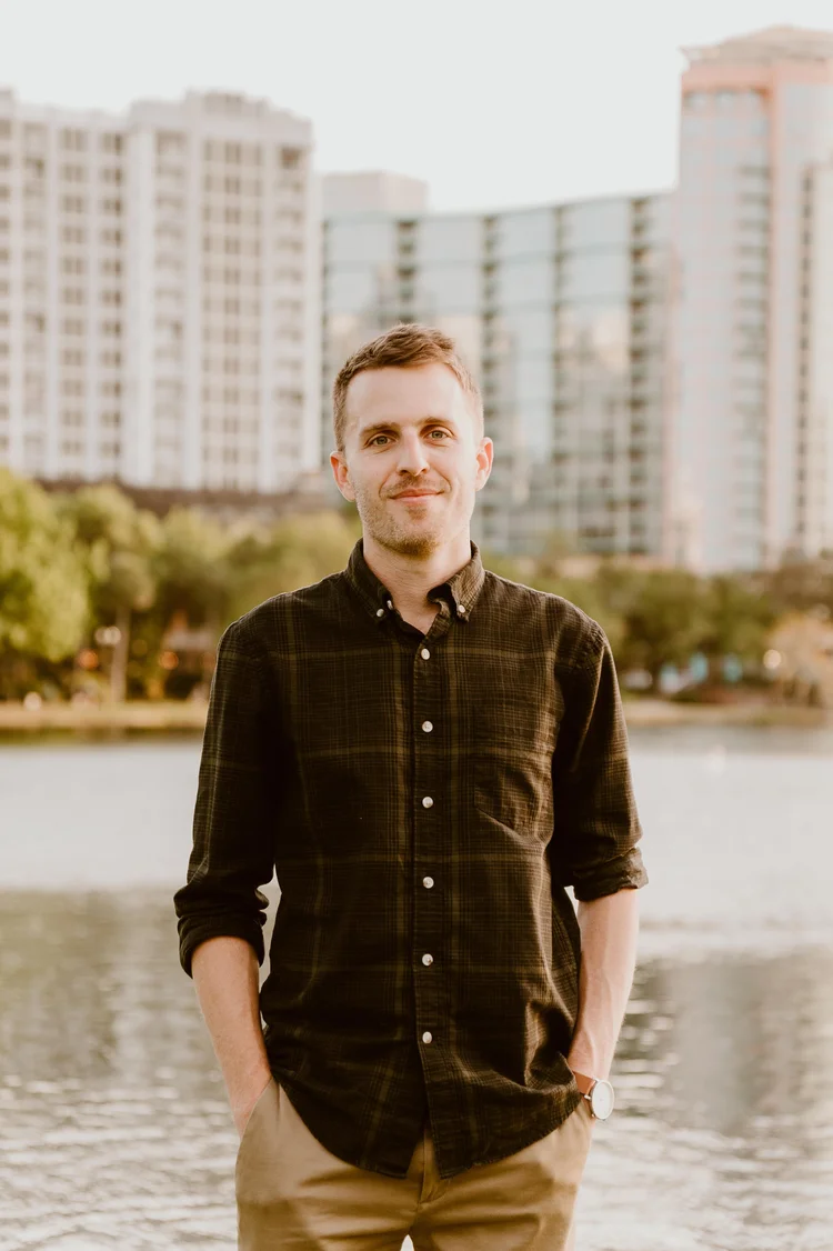 A young man stands outdoors near a body of water with tall modern buildings in the background, wearing a black button-up shirt and beige pants.