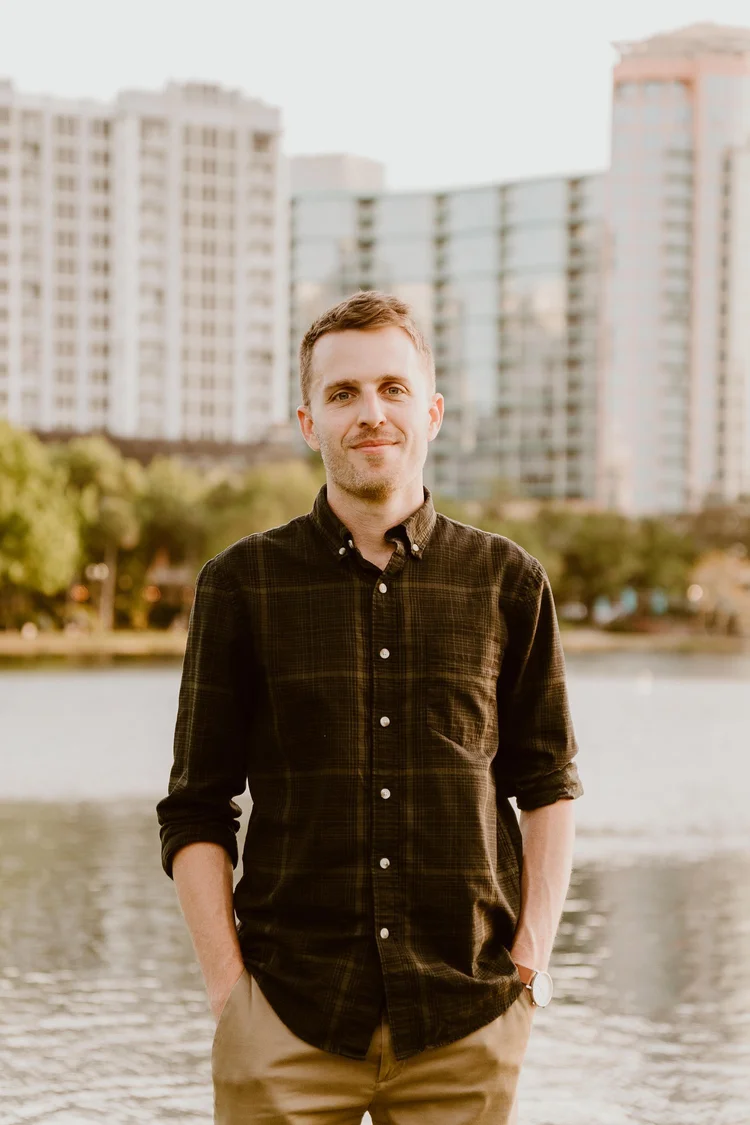 A young man with short brown hair, wearing a dark plaid button-up shirt and khaki pants, stands with hands in pockets by a body of water with city high-rise buildings in the background during sunset.